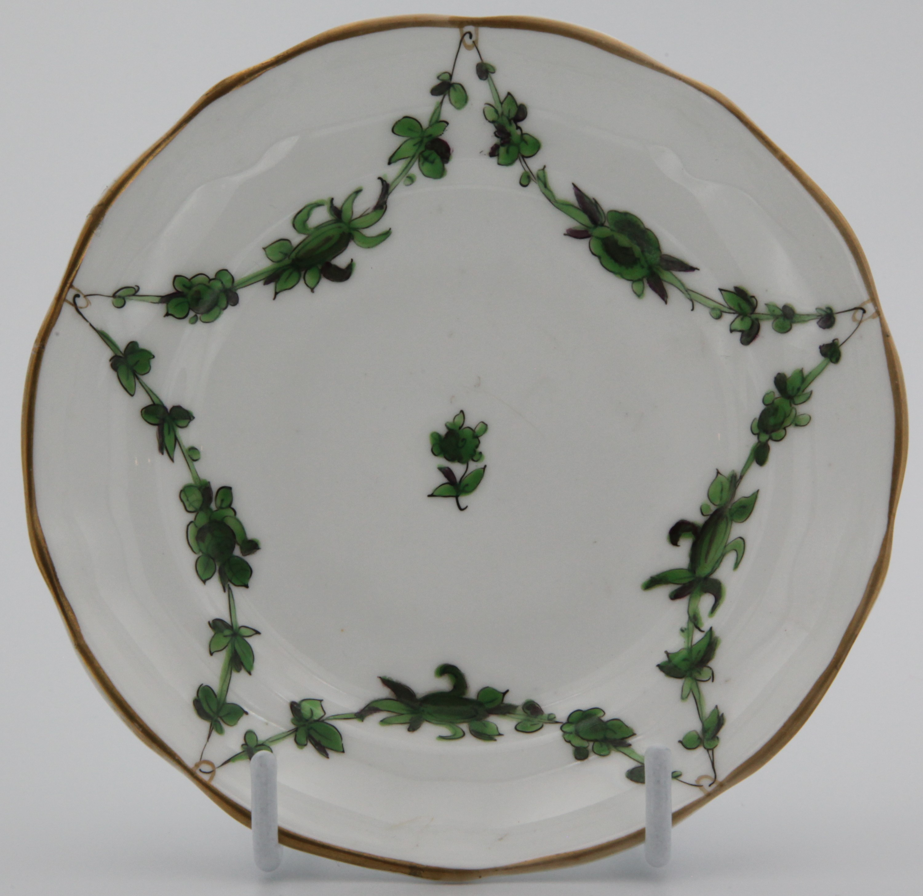 A Bristol-style saucer, of ogee and slightly lobed shape, painted in green monochrome with leaf and flower garlands suspended from gilt loops on gilt dentil rim, mark X 1798
