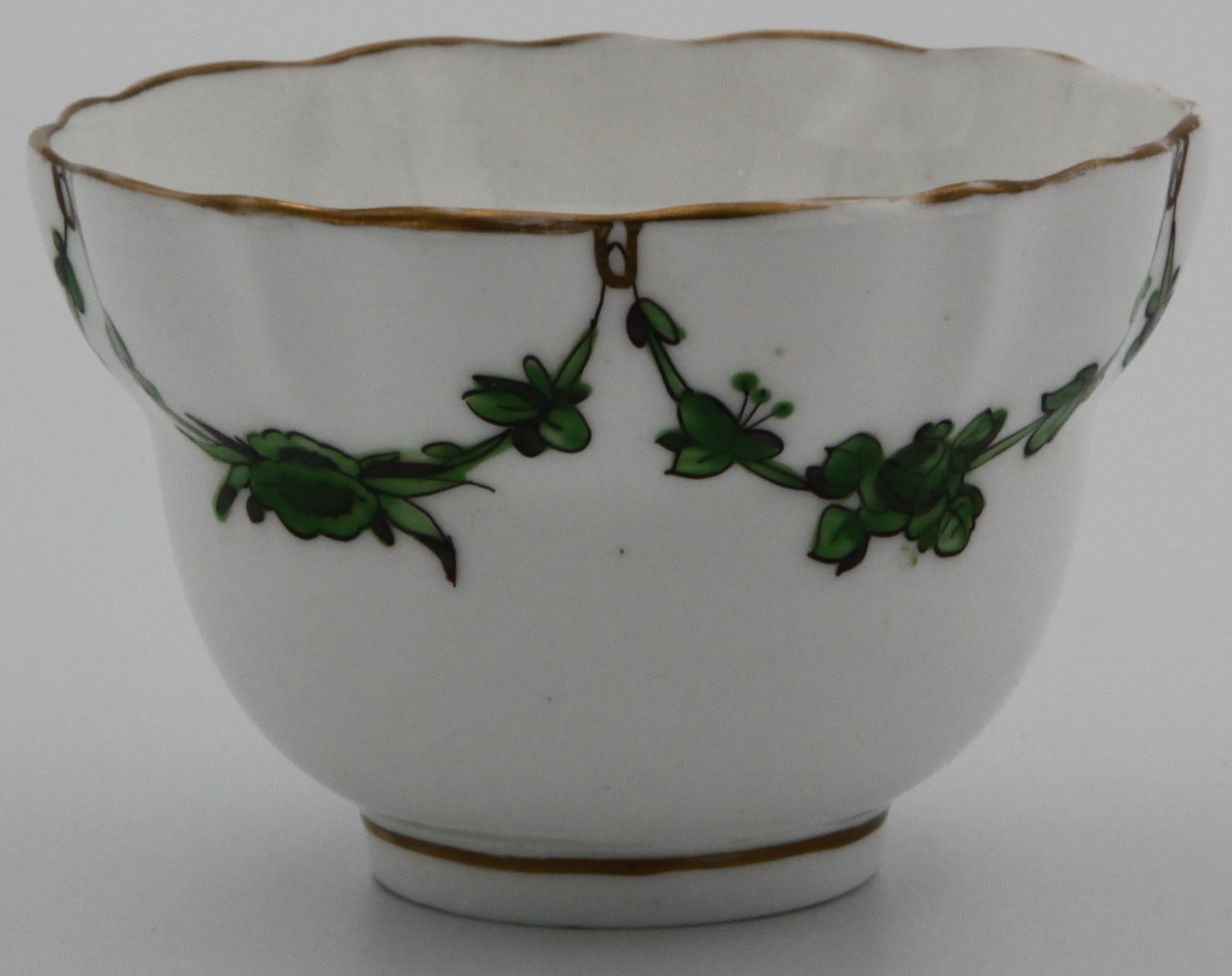 A Bristol-style tea bowl of ogee and slightly lobed shape, painted in green monochrome with leaf and flower garlands suspended from gilt loops on gilt dentil rim, mark X 1798