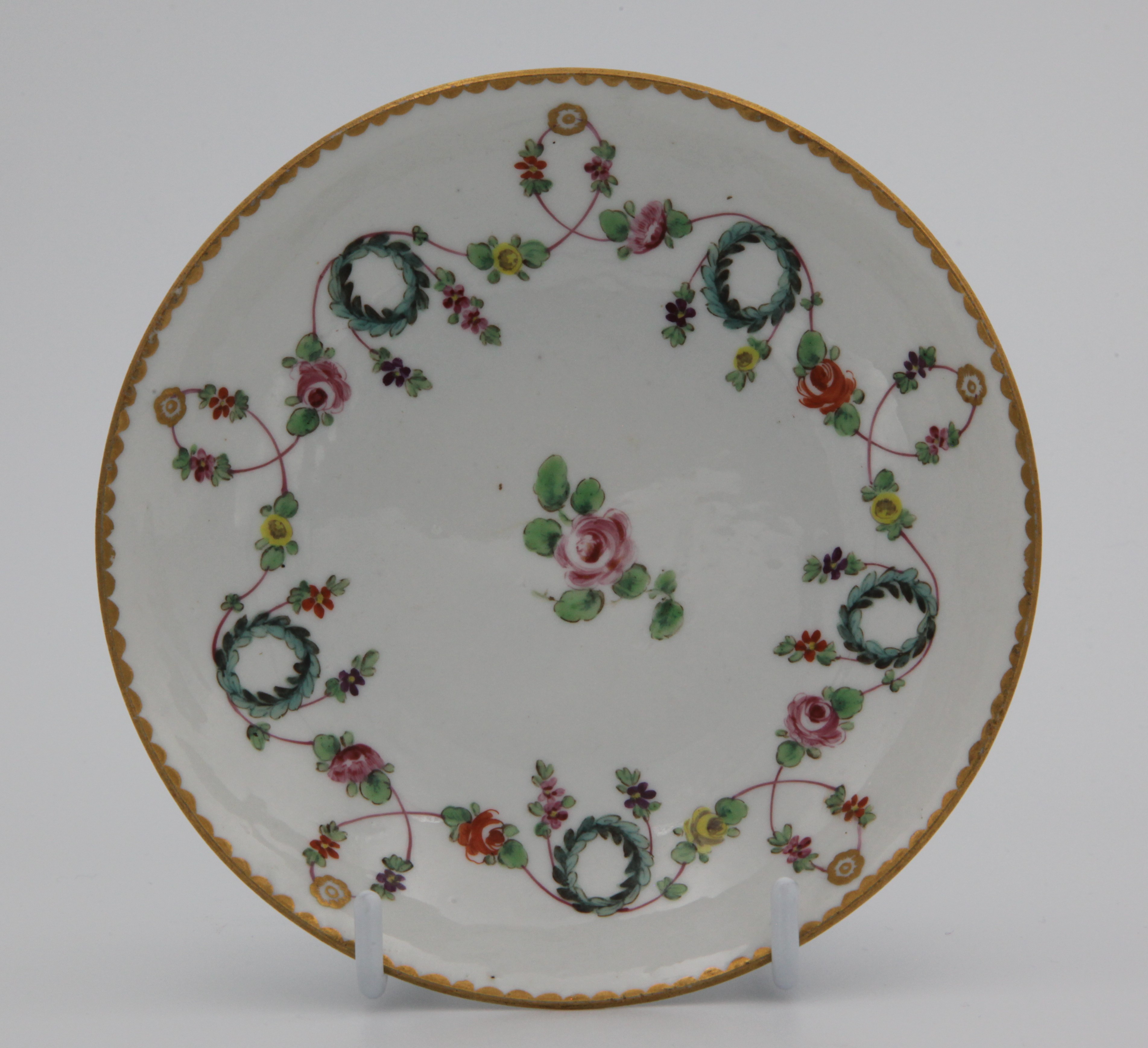 A very fine Bristol tea saucer, with rounded, slightly ogee sides, painted with colourful swags of leaves and flowers on a pink line, suspended from gilt roundels and looping over blue wreaths, with gilt dentil rim and central rose, 1. in gilt for Henry Bone
