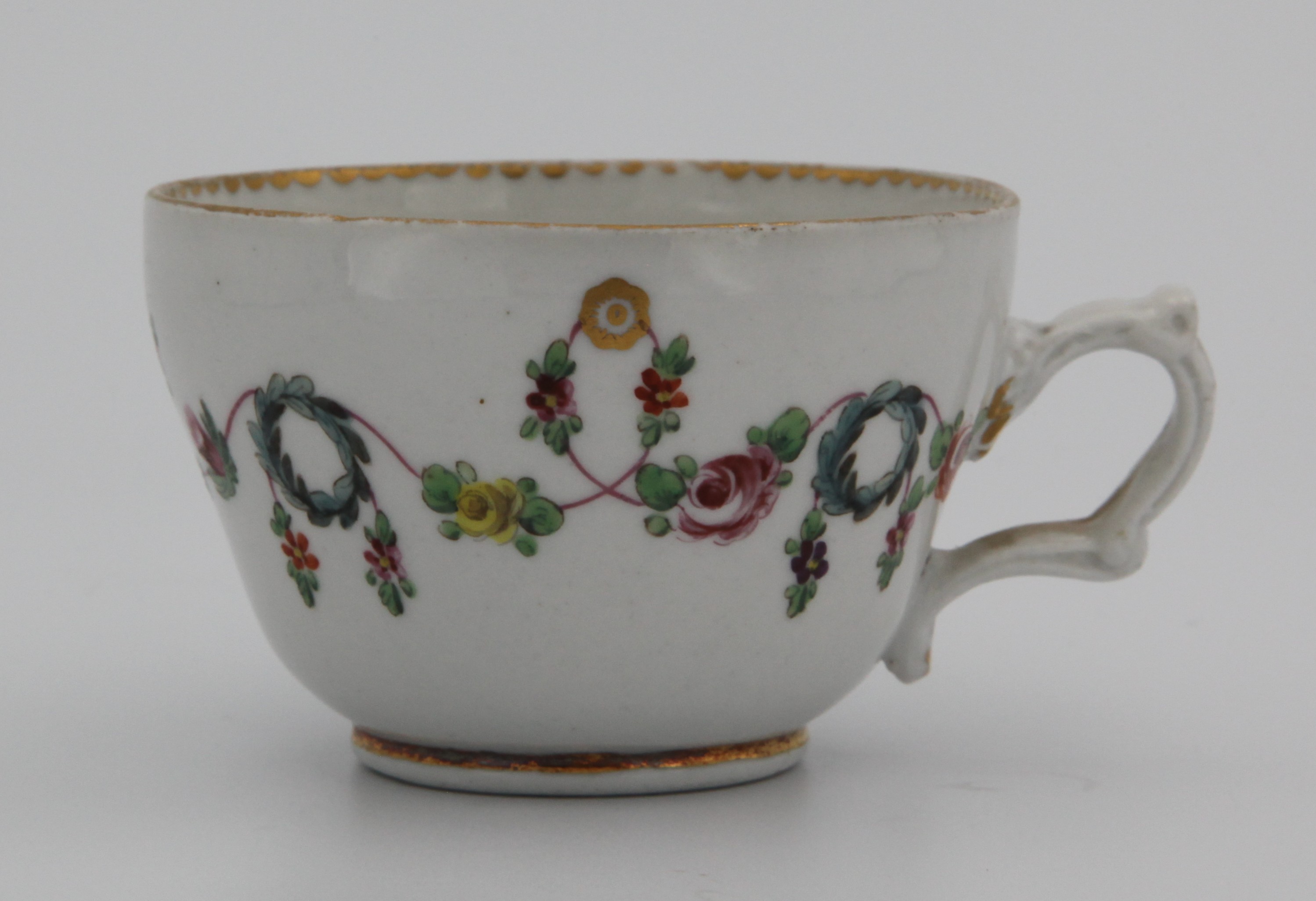 A very fine Bristol tea cup, slightly ogee, with elaborate moulded (but wonky) handle, painted with colourful swags of leaves and flowers on a pink line, suspended from gilt roundels and looping over blue wreaths, with gilt dentil rim and central rose in base of bowl, underglaze crossed swords, 1. in gilt for Henry Bone. Note slight firing mark decorated with gilt