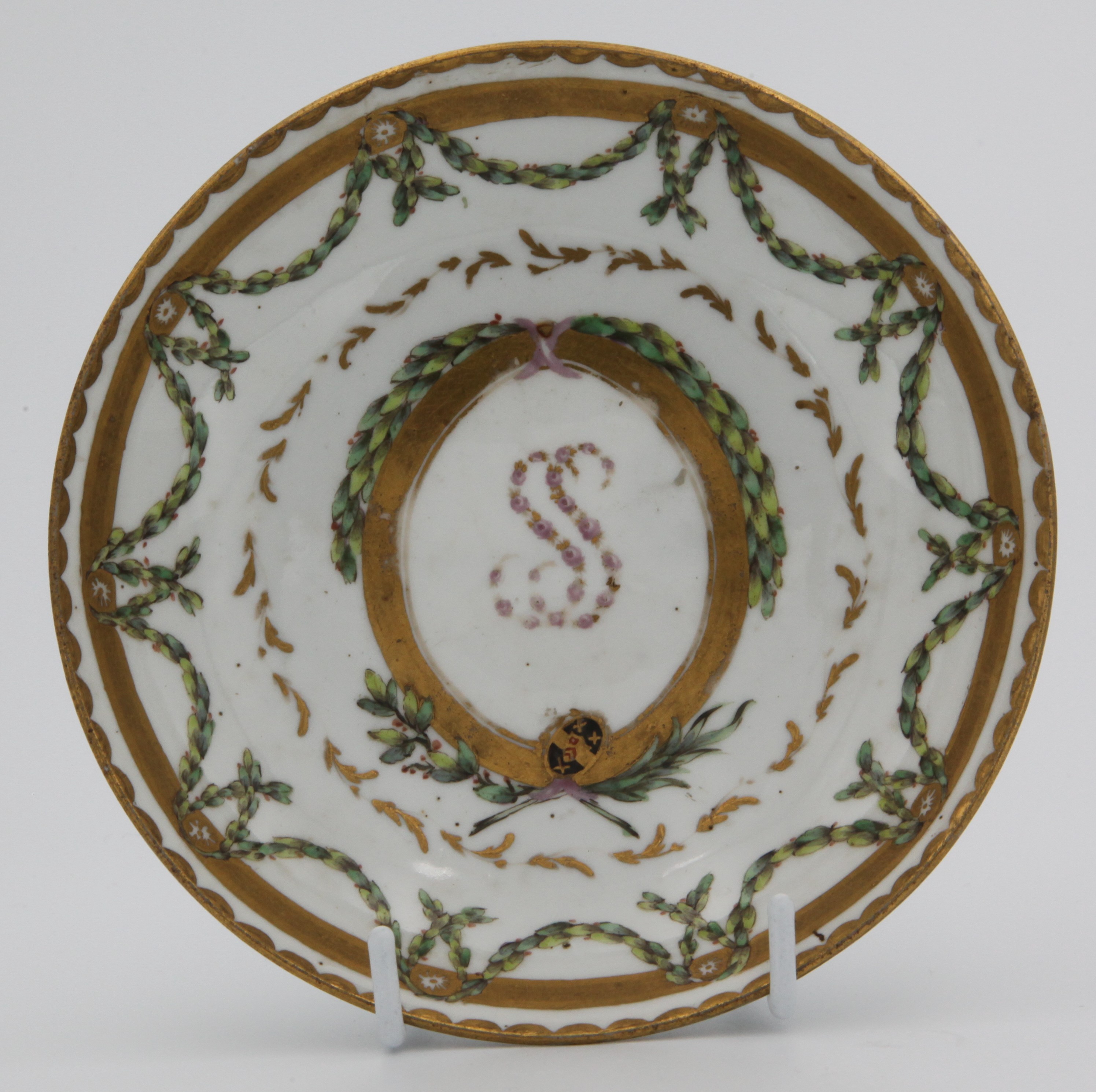 A Bristol coffee saucer from the Smith service, painted in enamels with green laurel garlands suspended from gilt roundels mounted on a thick gilt band, surrounding a ring of light gilt fronds in turn surrounding a central gilt-framed panel, adorned with laurel garlands and pink ribbons and containing both the arms of Smith in black, red and gilt and the initials SS (Sarah Smith) in tiny pink and gilt flowers, all surrounded by a gilt dentil rim. circa 1774-75. Mark: blue X