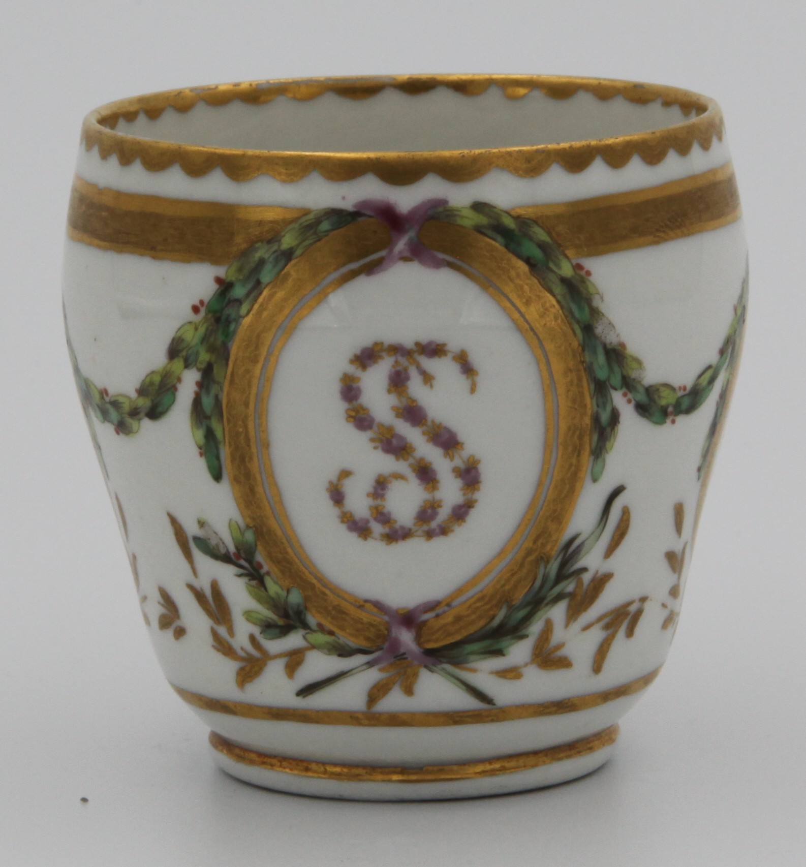 A Bristol coffee cup from the Smith service, of ogee shape, with angular, gilt-decorated handle, painted in enamels with green laurel garlands resting on oval gilt panels, two containing the arms of Smith in black, red and gilt and the larger panel containing the initials SS (Sarah Smith) in tiny pink and gilt garlands and all three panelse resting on gilt fronds, all between heavy gilt lines and under gilt dentil rim, circa 1774-75. Mark: blue X