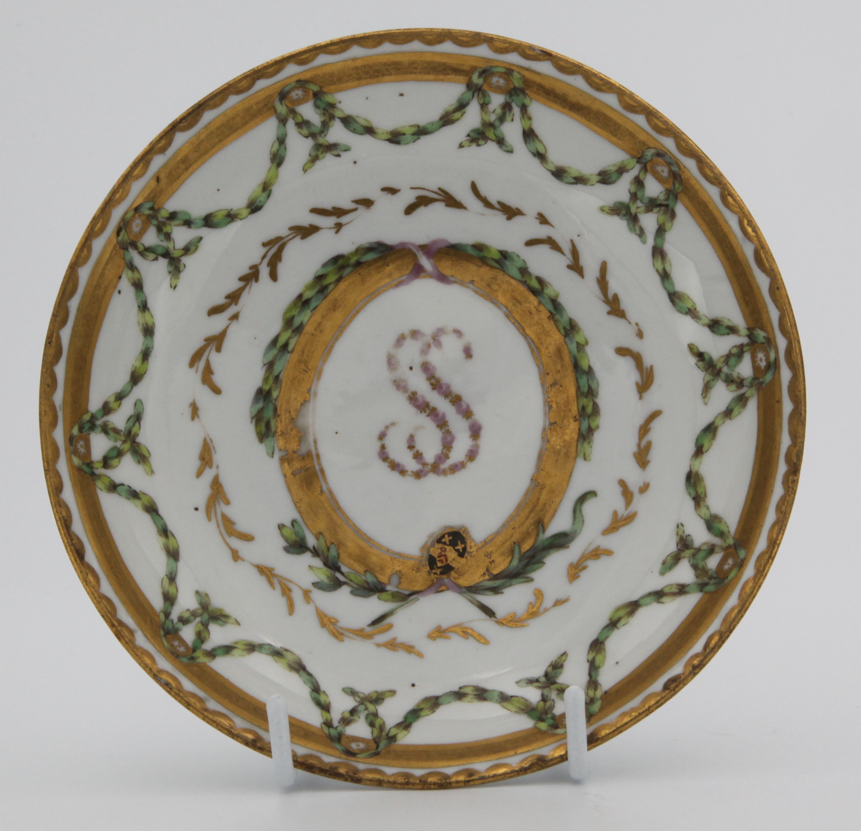 A Bristol sucrier stand from the Smith service, painted in enamels with green laurel garlands suspended from gilt roundels mounted on a thick gilt band, surrounding a ring of light gilt fronds in turn surrounding a central gilt-framed panel, adorned with laurel garlands and pink ribbons and containing both the arms of Smith in black, red and gilt and the initials SS (Sarah Smith) in tiny pink and gilt flowers, all surrounded by a gilt dentil rim. circa 1774-75. Mark: grey X