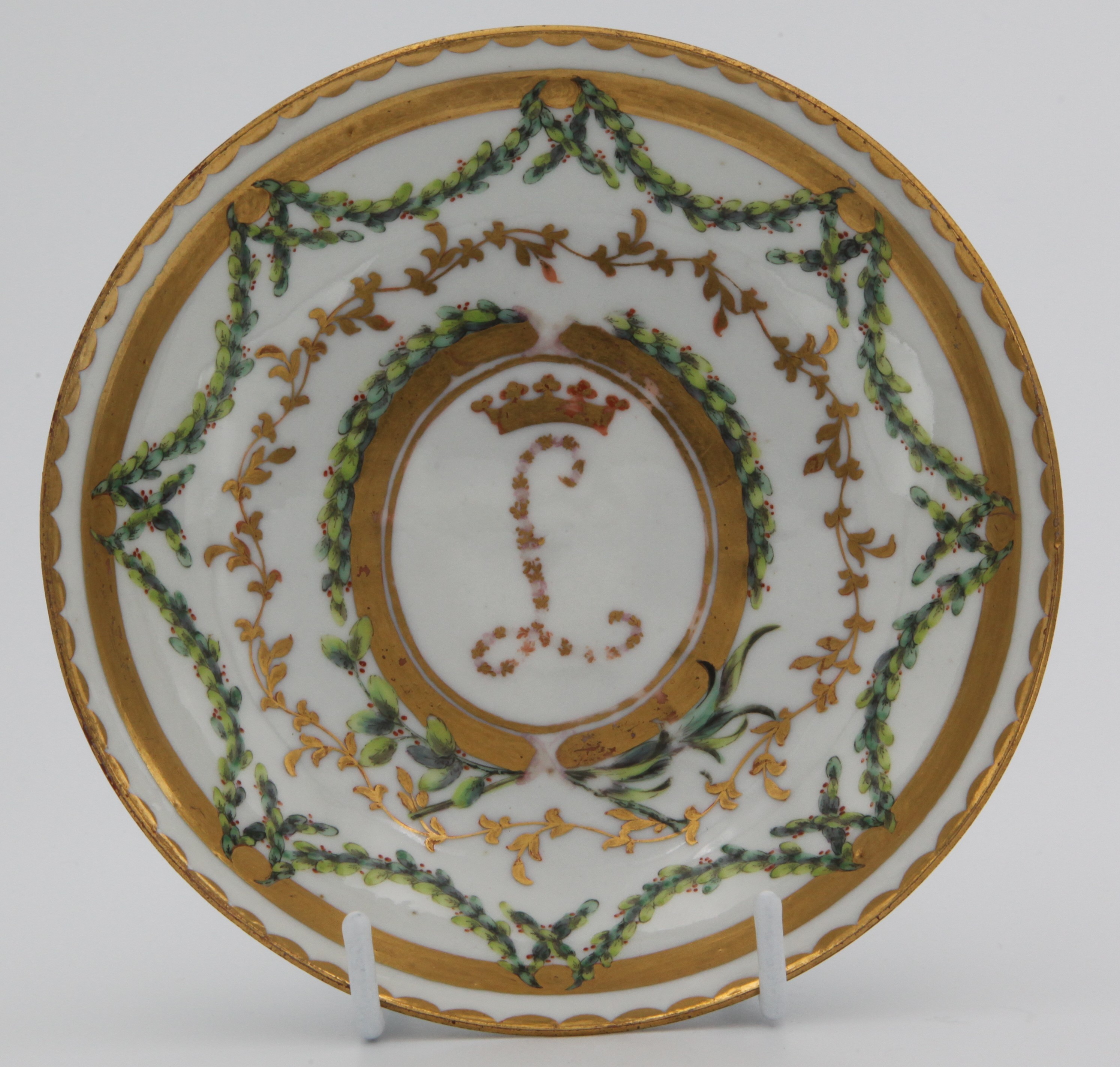 A rare Bristol ogee-shaped coffee saucer, from the Leinster service, decorated in coloured flower heads with the intial L beneath a gilt ducal coronet, within oval gilt panels, the cup with two grisaille head and shoulder studies of young men, united by green and iron red laurel swags, gilt line, leaf scroll and dentil borders, circa 1775, blue painted X and numeral 3 in gilt.
