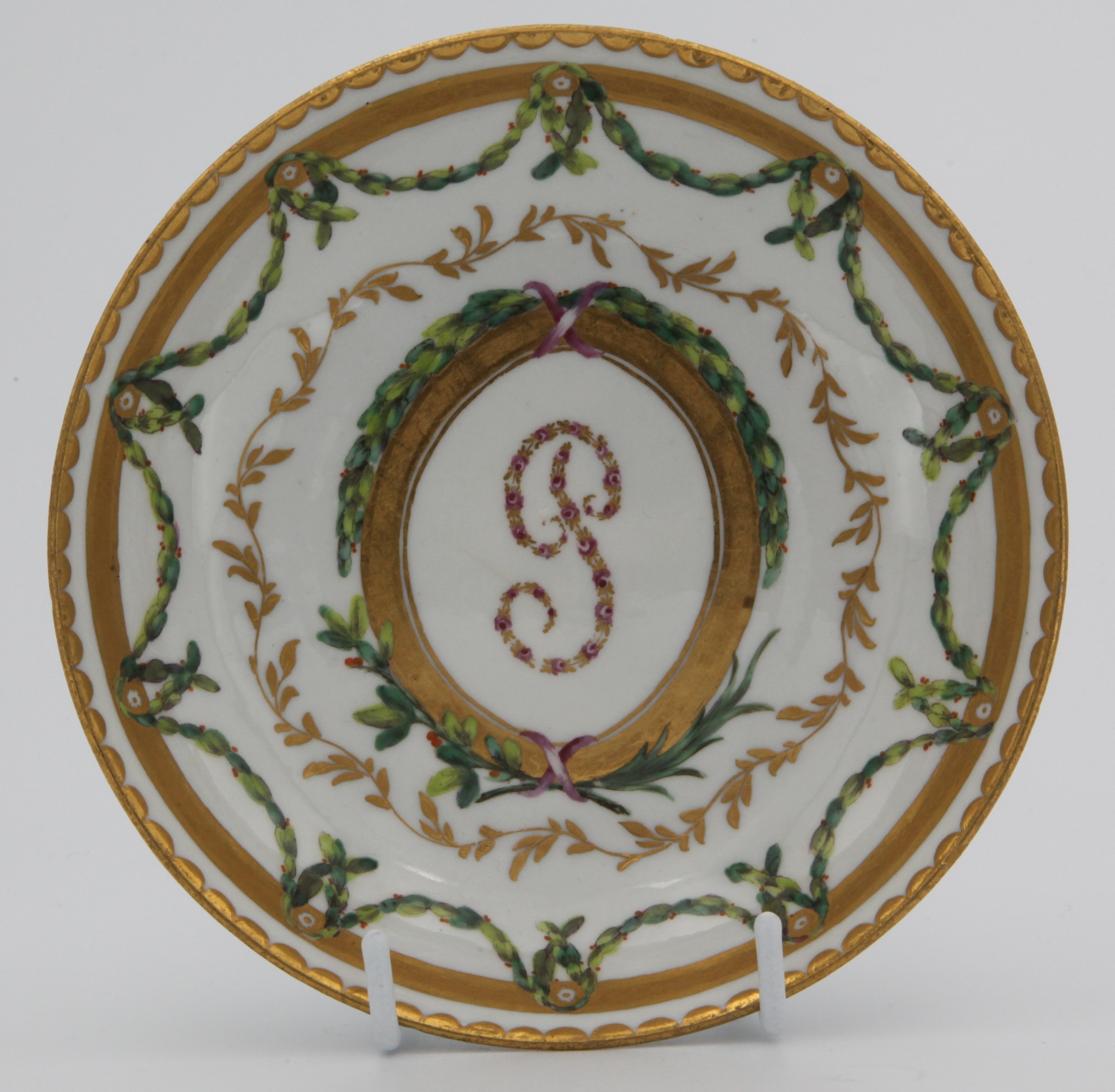 A Bristol tea saucer, from the Plumer Service, with initial P in pink roses and gilt, within a tooled oval gilt panel, and with grisaille portrait medallions, within green, iron red and gilt laurel and leaf scroll borders, gilt dentil rims, circa 1774-75, blue painted X marks. See F S Mackenna, Plymouth and Bristol Porcelain, number 29, for the sucrier and cover from this service
