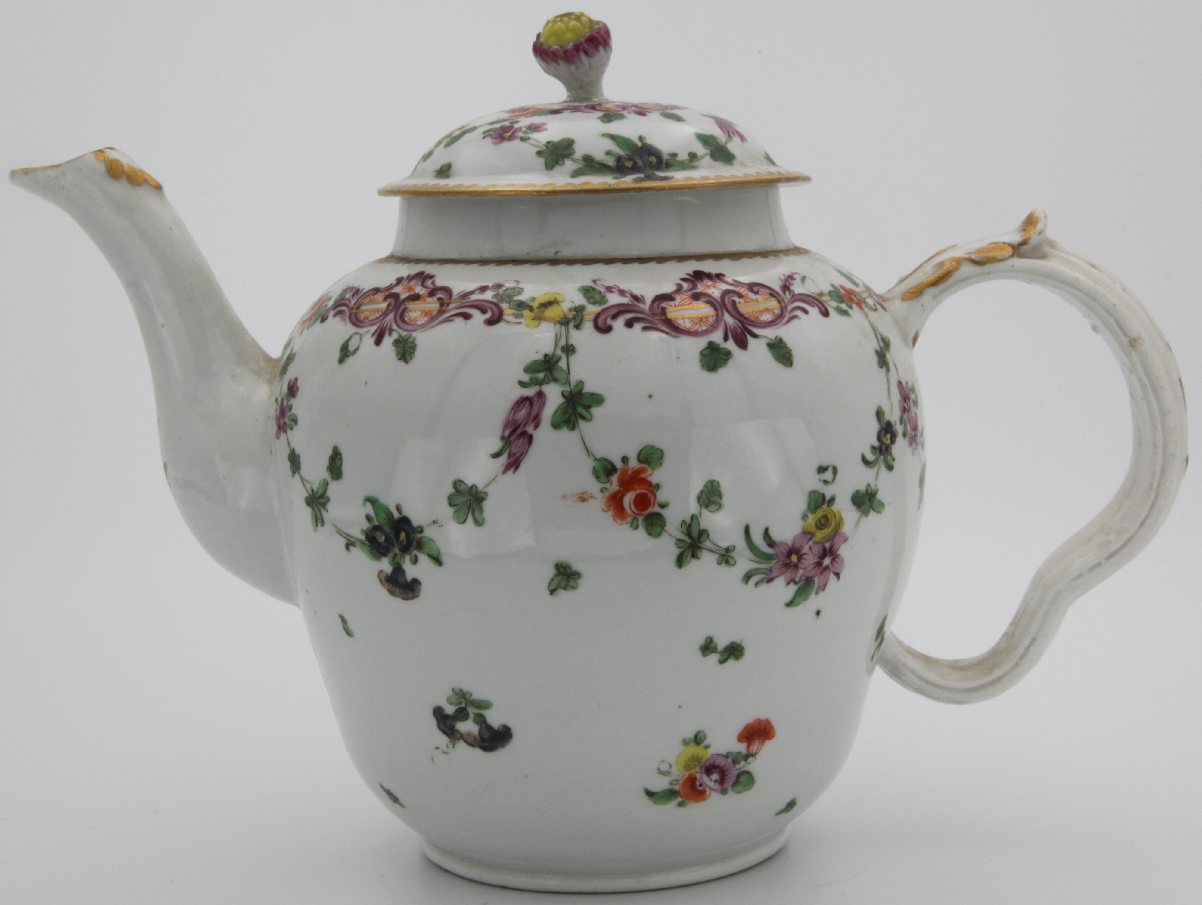 A Bristol porcelain teapot with ear-shaped moulded and gilt decorated handle, and cover with purple bud finial, painted in generally green and purple enamels with flower sprigs and garlands suspended from a floral and leafy scroll border, and with scattered leaves and flowers, under a gilt dentil rim. c. 1775. No mark.