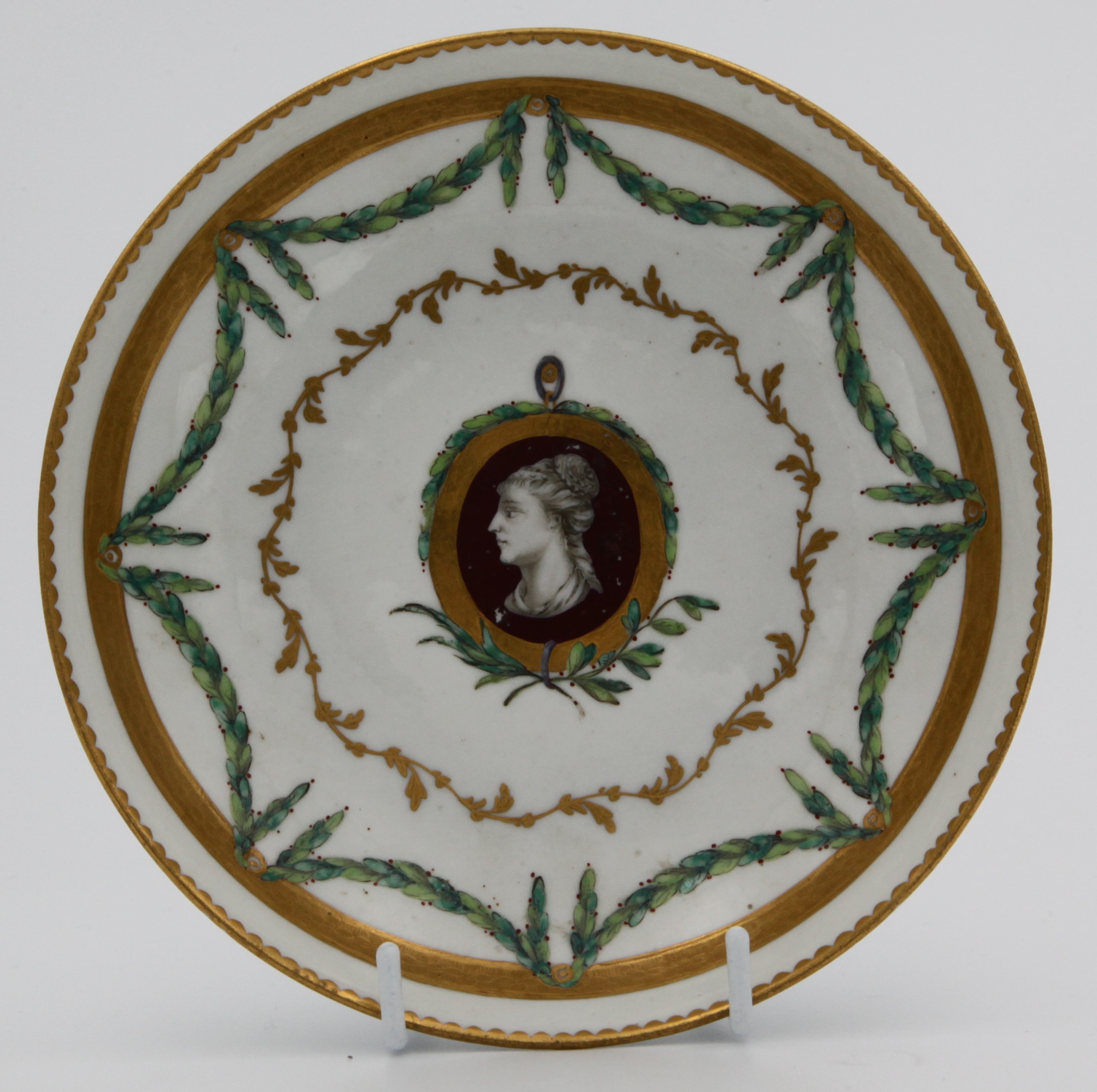 A Bristol 'classical portraits' chocolate cup stand, painted with classical heads within berried green wreaths and borders of gilt foliage and berried green garlands, gilt dentil rims, circa 1775-80. No mark