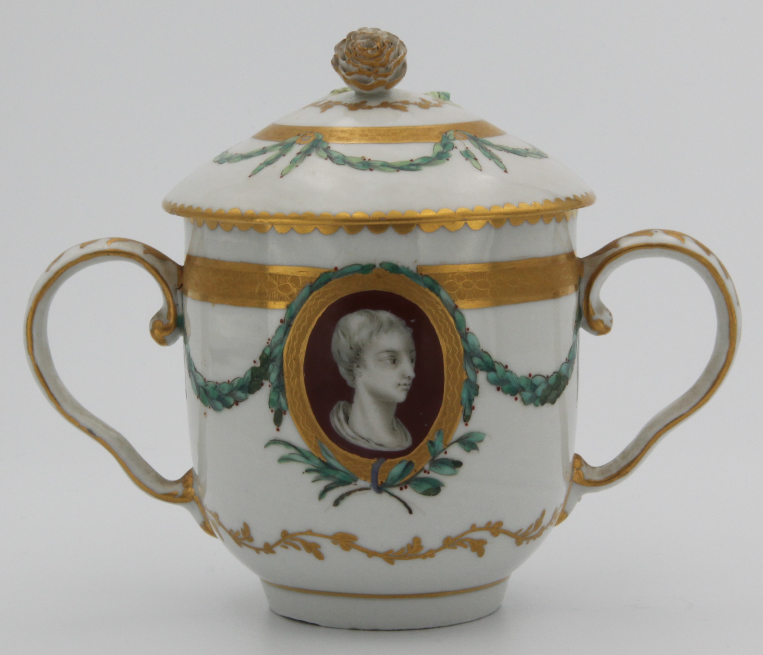 A Bristol 'classical portraits' twin handled chocolate cup and cover with bud finial, painted with classical heads within berried green wreaths and borders of gilt foliage and berried green garlands, gilt dentil rims, circa 1775-80. No mark