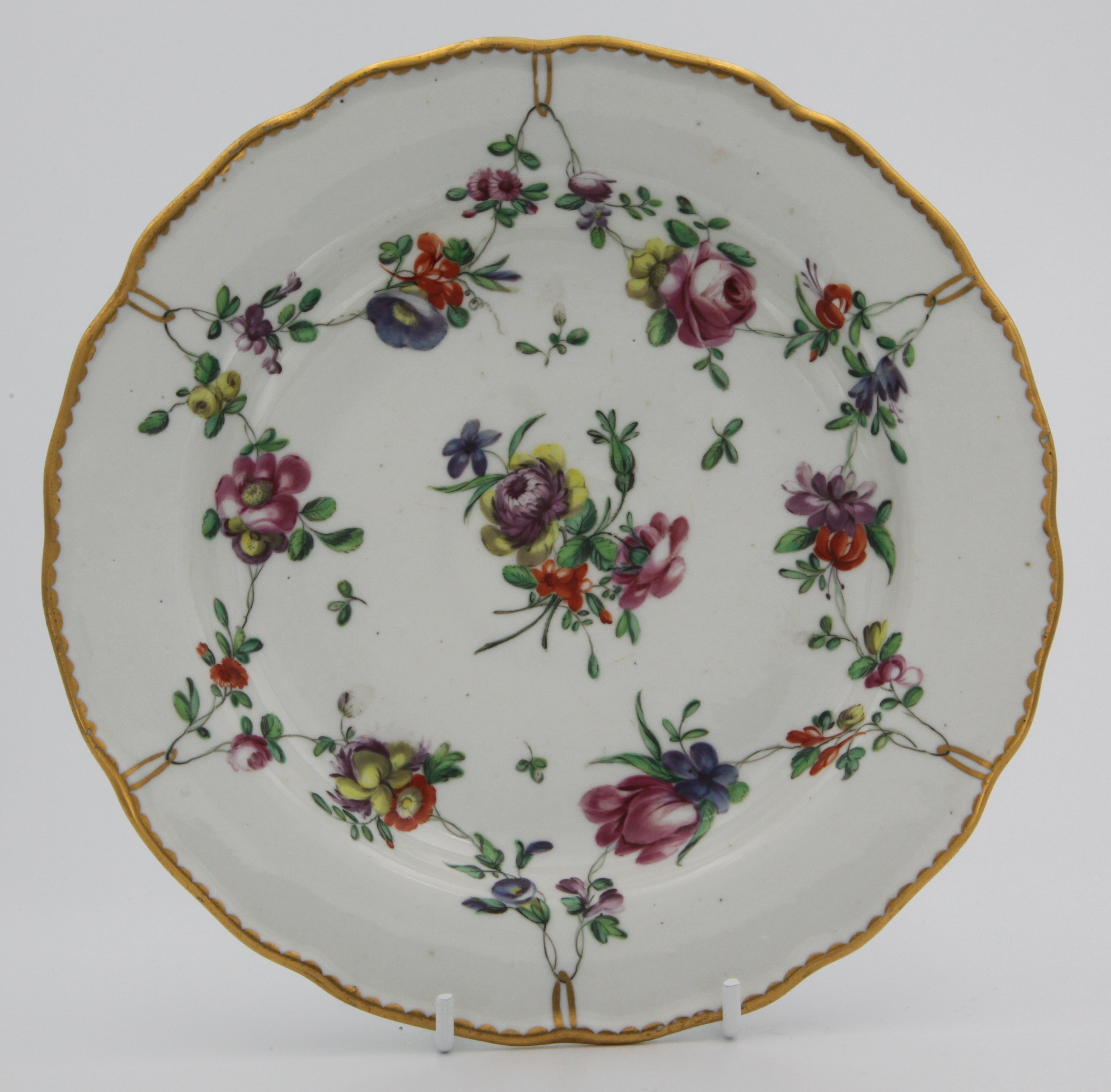 A fine Bristol plate painted with garlands of flowers suspended from gilt rings around a central bouquet, gilt dentil rim, blue Xb mark, circa 1775-80. Double reinforcement ring to base.