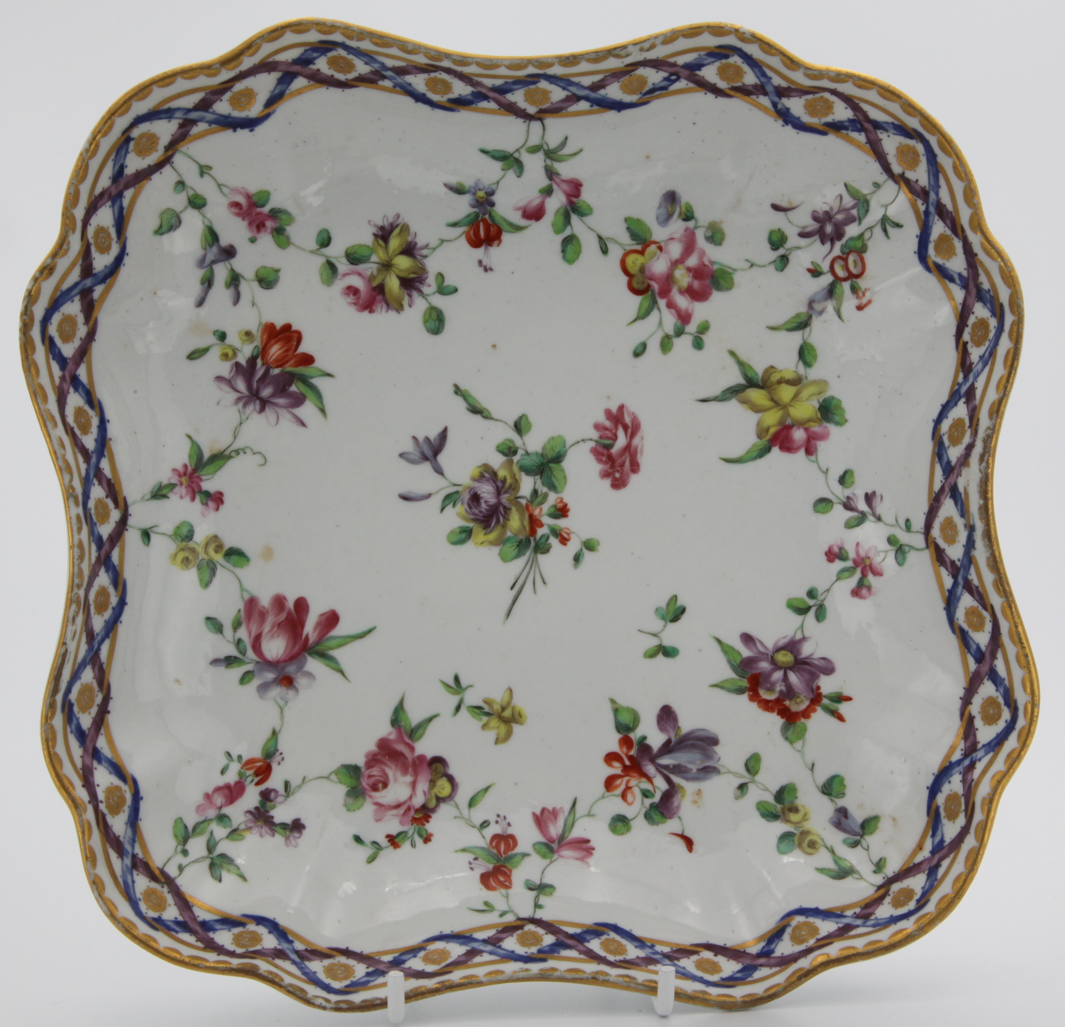 A Bristol lobed square dessert dish painted in the Sevres manner with the 'double ribbon' pattern of entwined ribbons supporting garlands of flowers and foliage, gilt dentil rim, blue X mark, circa 1775-80, 21cm.