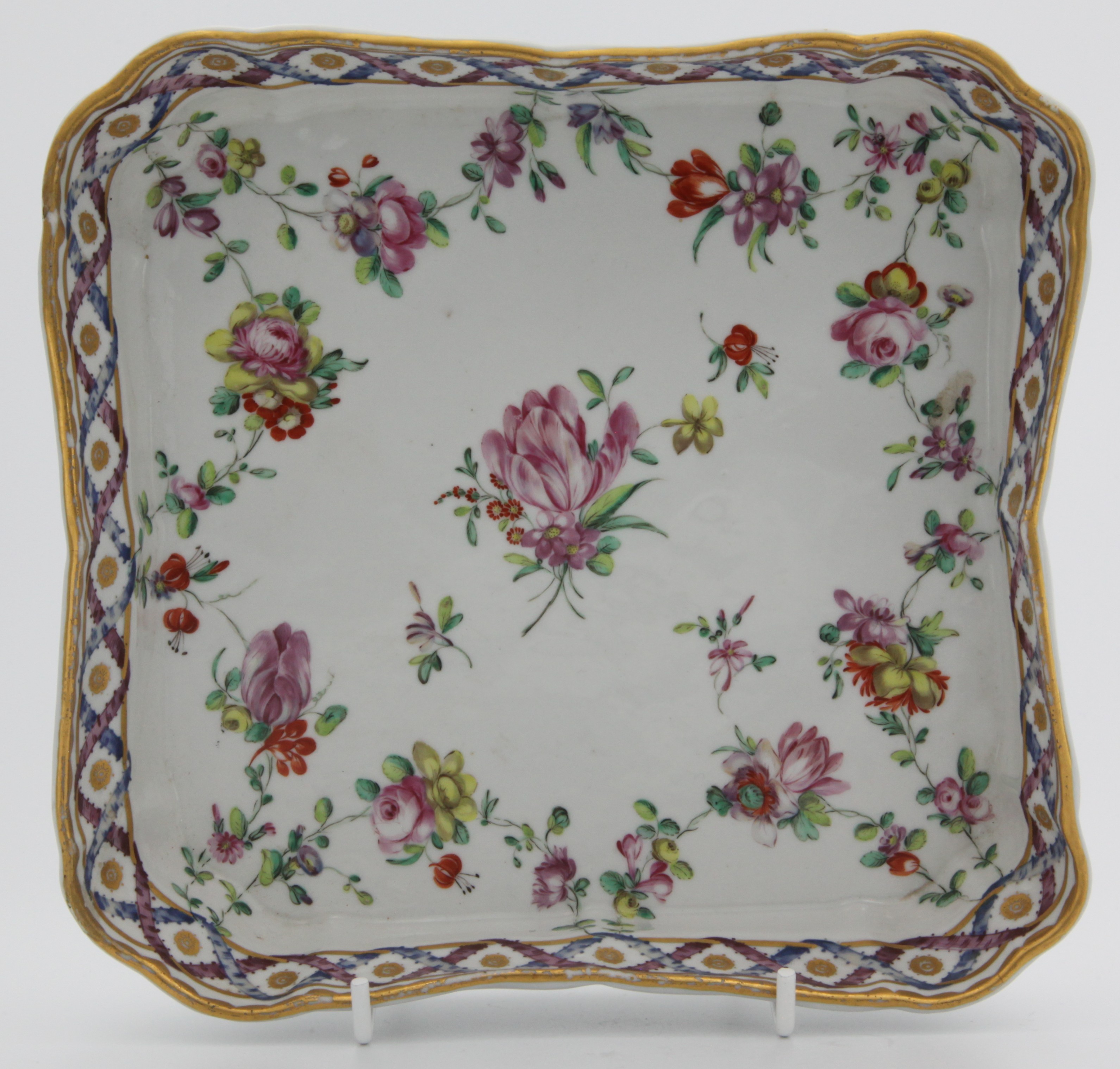 A Bristol square shaped dish painted in the 'double ribbon' pattern of flower sprays and garlands suspended from entwined blue and puce ribbons, gilt dentil rim, blue X mark with gilt b., circa 1775-8.