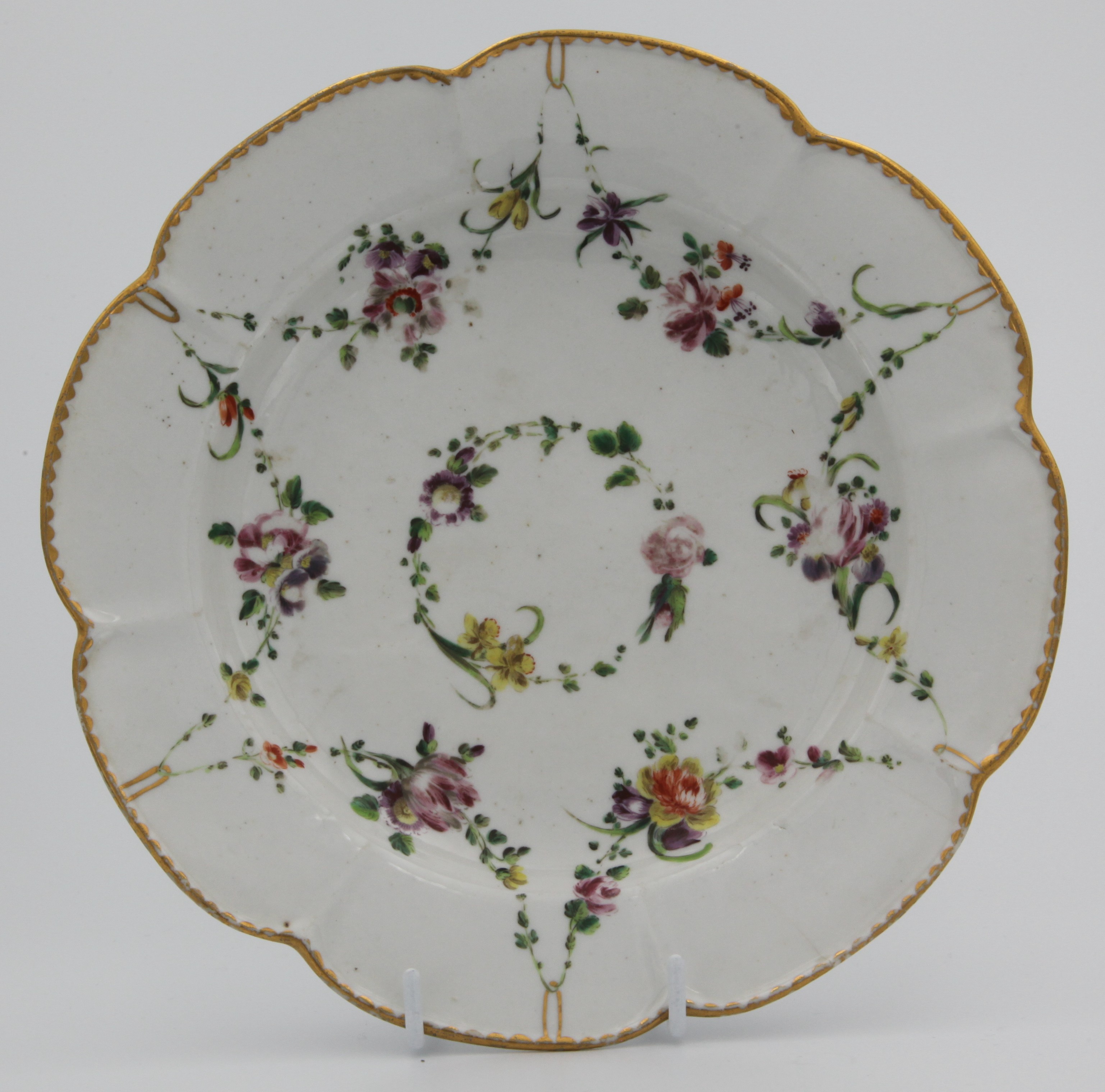 A Bristol lobed porcelain plate, crudely painted swags of flowers suspended from gilt loops hanging from gilt dentil rim. Central ring of flowers and leaves.  c.1775. Grey X mark. Double reinfocement ring to base