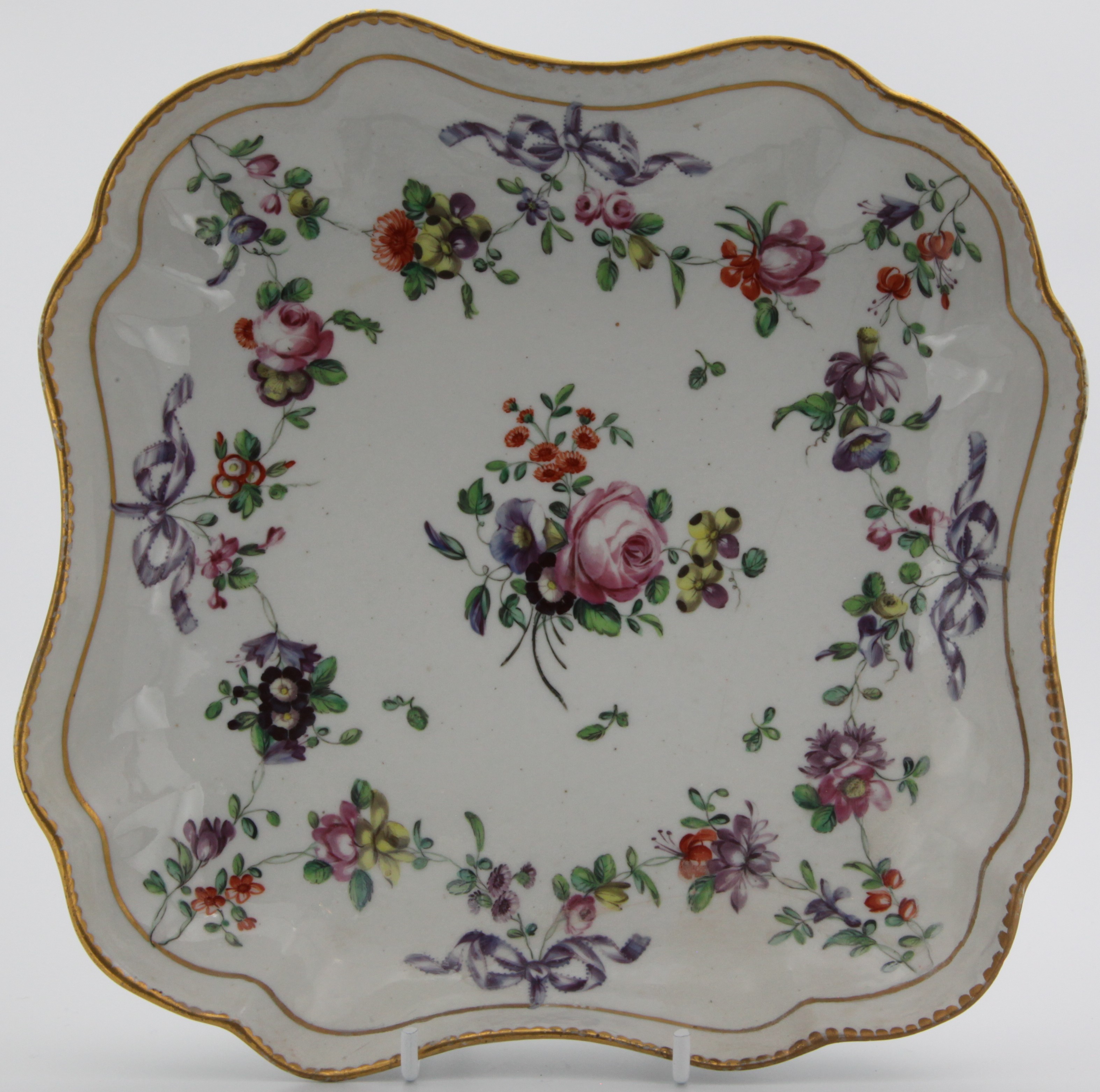 A Bristol square dish, painted in enamels with a colouful floral garland suspended from a gilt band and from purple ribbons, with central floral spray, all within gilt dentil rims. Blue Xb mark