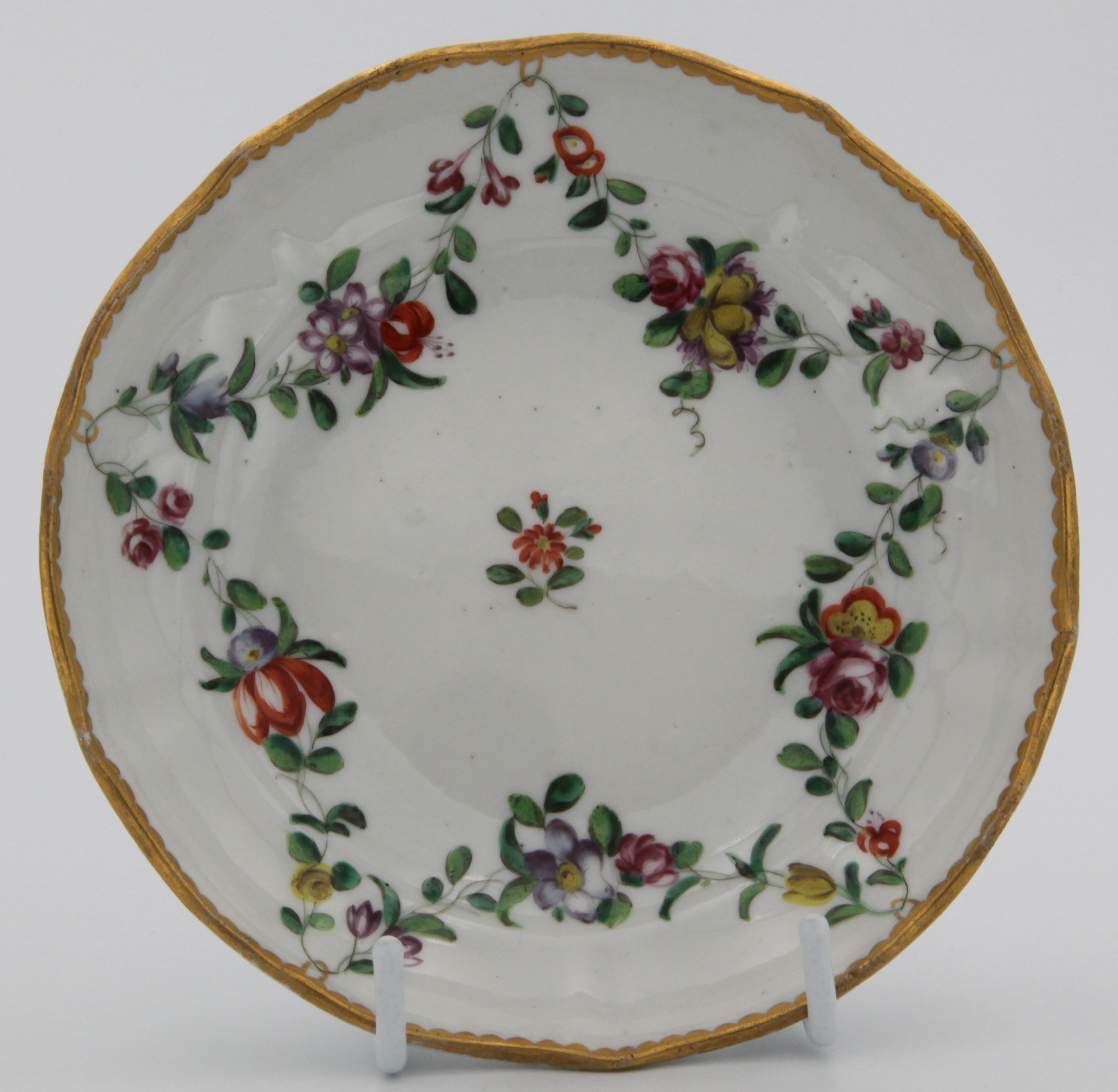 Bristol saucer painted with chains of flowers hanging from gold loops around a central red flower spray and the loops suspended from a heavy gilt dentil rim. c. 1775-80 Mark: blue X3.