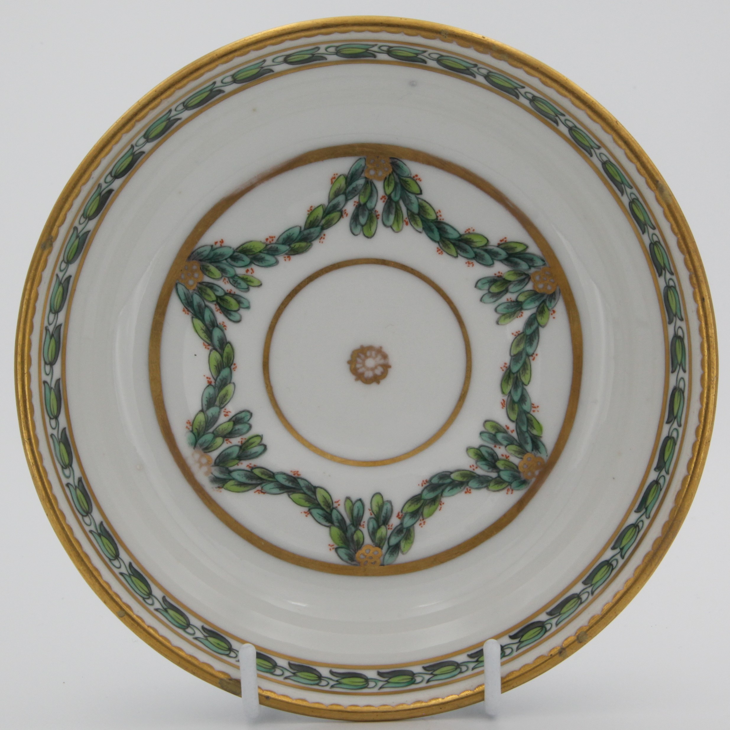 A Bristol coffee saucer, painted in green and iron red with laurel swags, suspended from gilt bands within green leaf scroll bands and gilt dentil rims, circa 1775. Blue X1 mark
