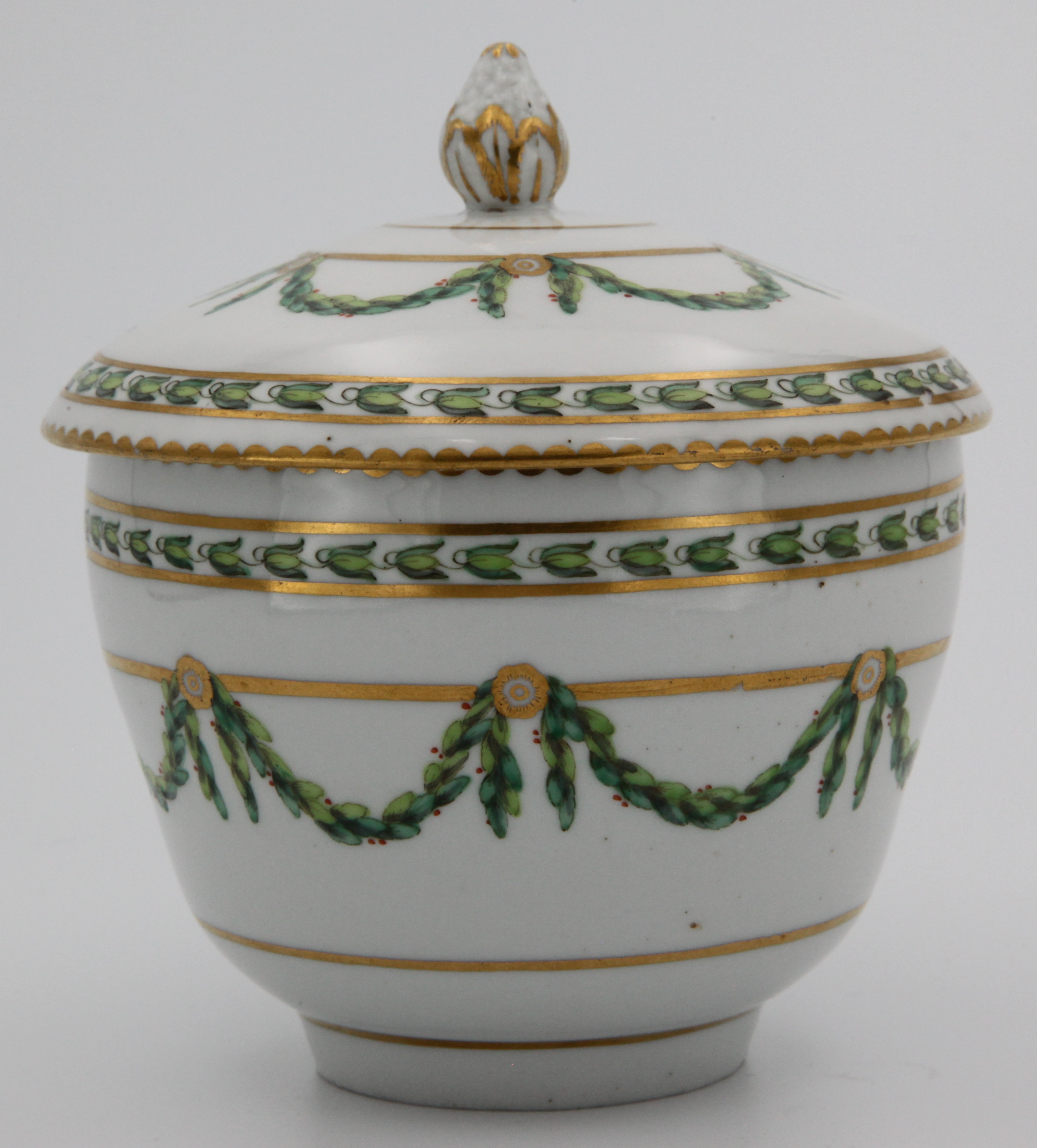 A Bristol ogee shaped sucrier and cover, with pinecone knop, painted in green and iron red with laurel swags, suspended from gilt flower head roundels and bands, within stiff leaf and gilt dentil borders, circa 1775, blue painted X mark.