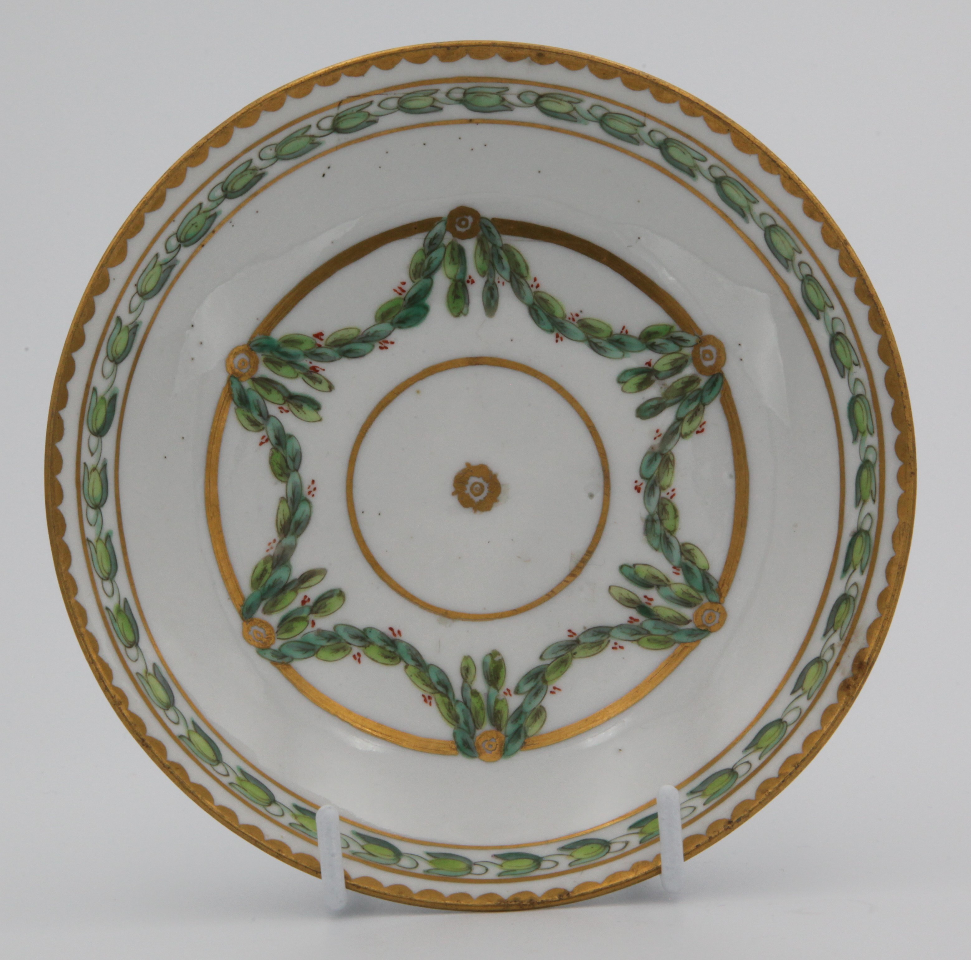A Bristol round-shaped saucer, painted in green and iron red with laurel swags, suspended from gilt bands within green leaf scroll bands and gilt dentil rims, circa 1775, blue painted X1. mark.