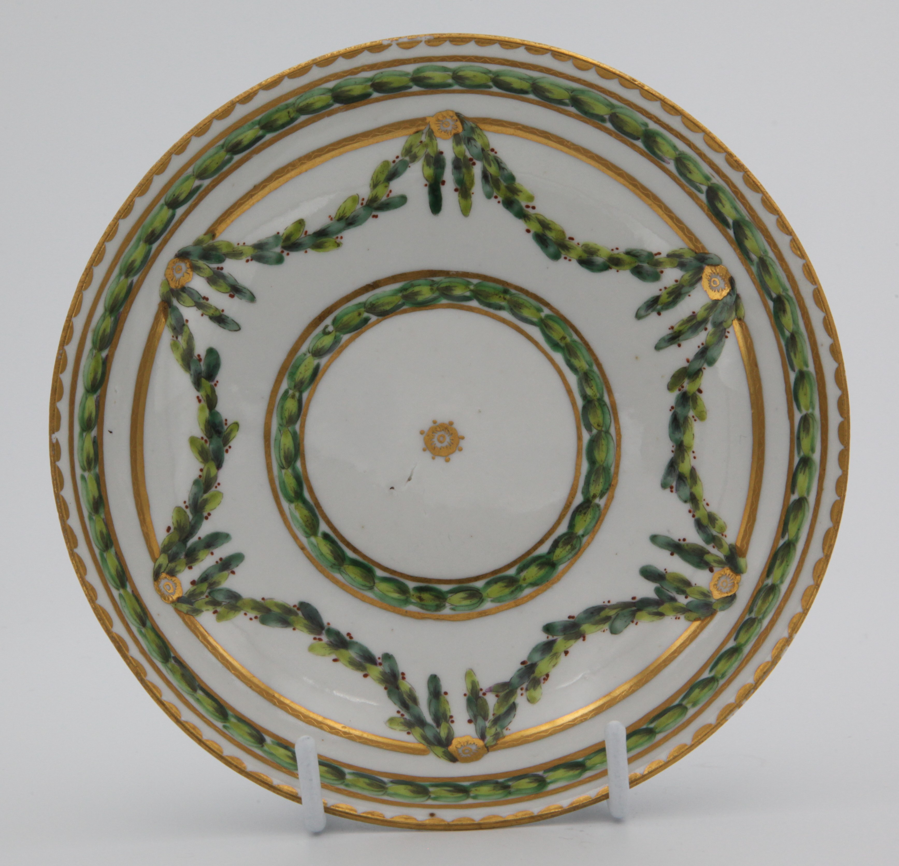 A Bristol tea saucer, painted in green iron red and gilt with laurel garlands, suspended from roundels hanging on gilt bands, within gilt line and dentil borders, circa 1772-75, blue painted X mark.