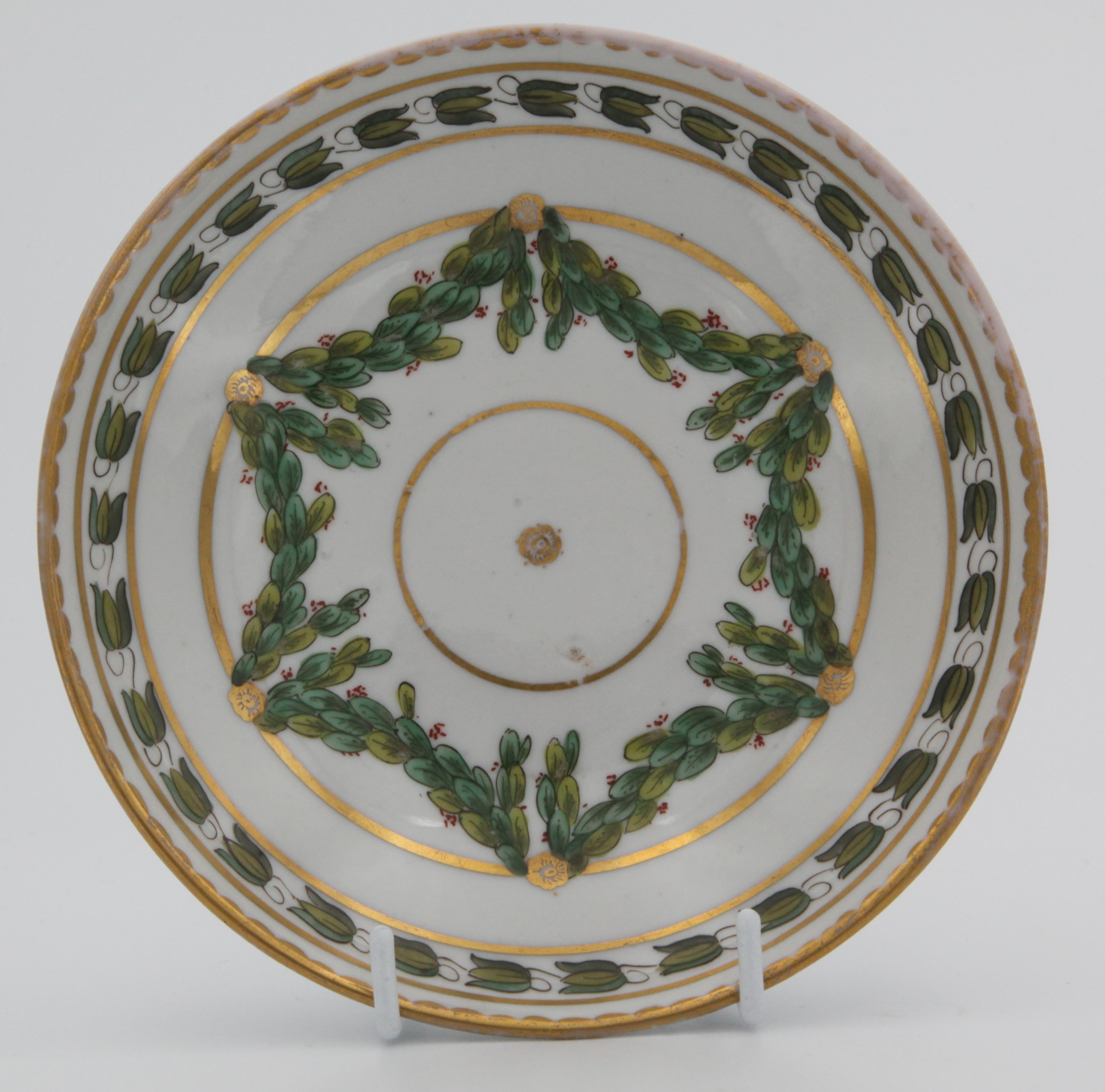A Bristol coffee saucer, painted in green and iron red with laurel swags, suspended from gilt bands within green leaf scroll bands and gilt dentil rims, circa 1775. Blue X1 mark