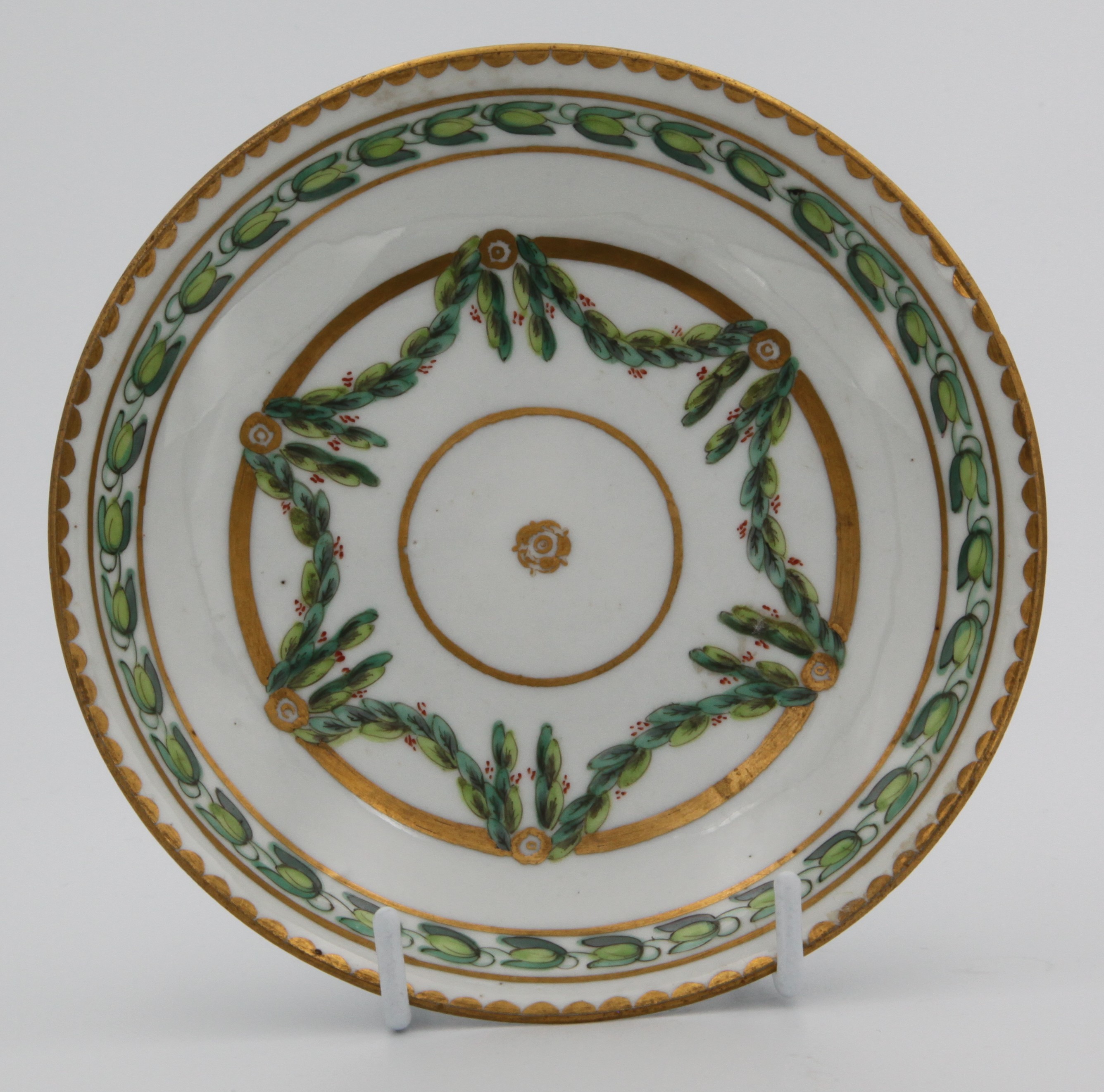 A Bristol coffee saucer, painted in green and iron red with laurel swags, suspended from gilt bands within green leaf scroll bands and gilt dentil rims, circa 1775. Blue X1. mark