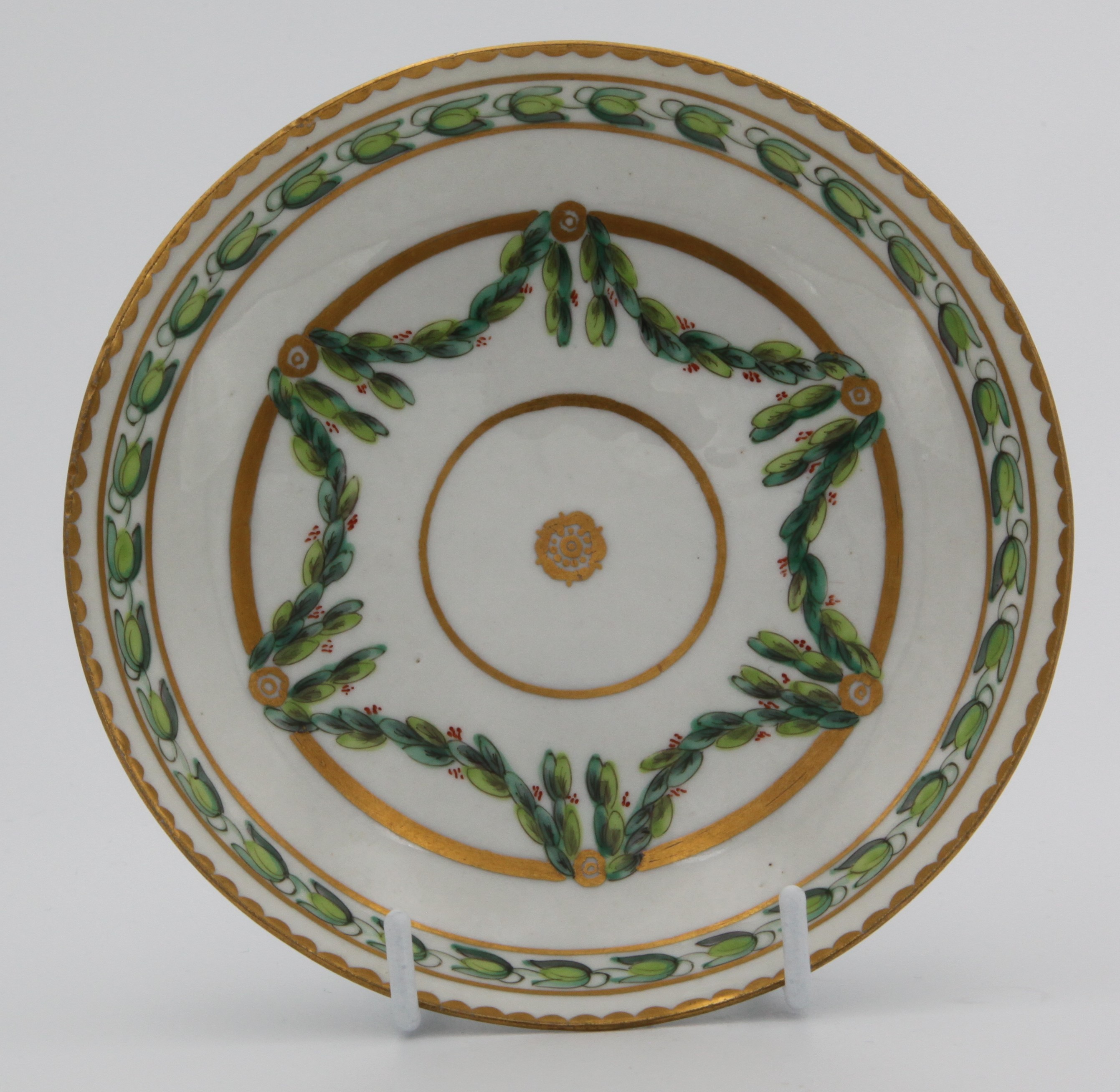 A Bristol coffee saucer, painted in green and iron red with laurel swags, suspended from gilt bands within green leaf scroll bands and gilt dentil rims, circa 1775. Blue X1. mark