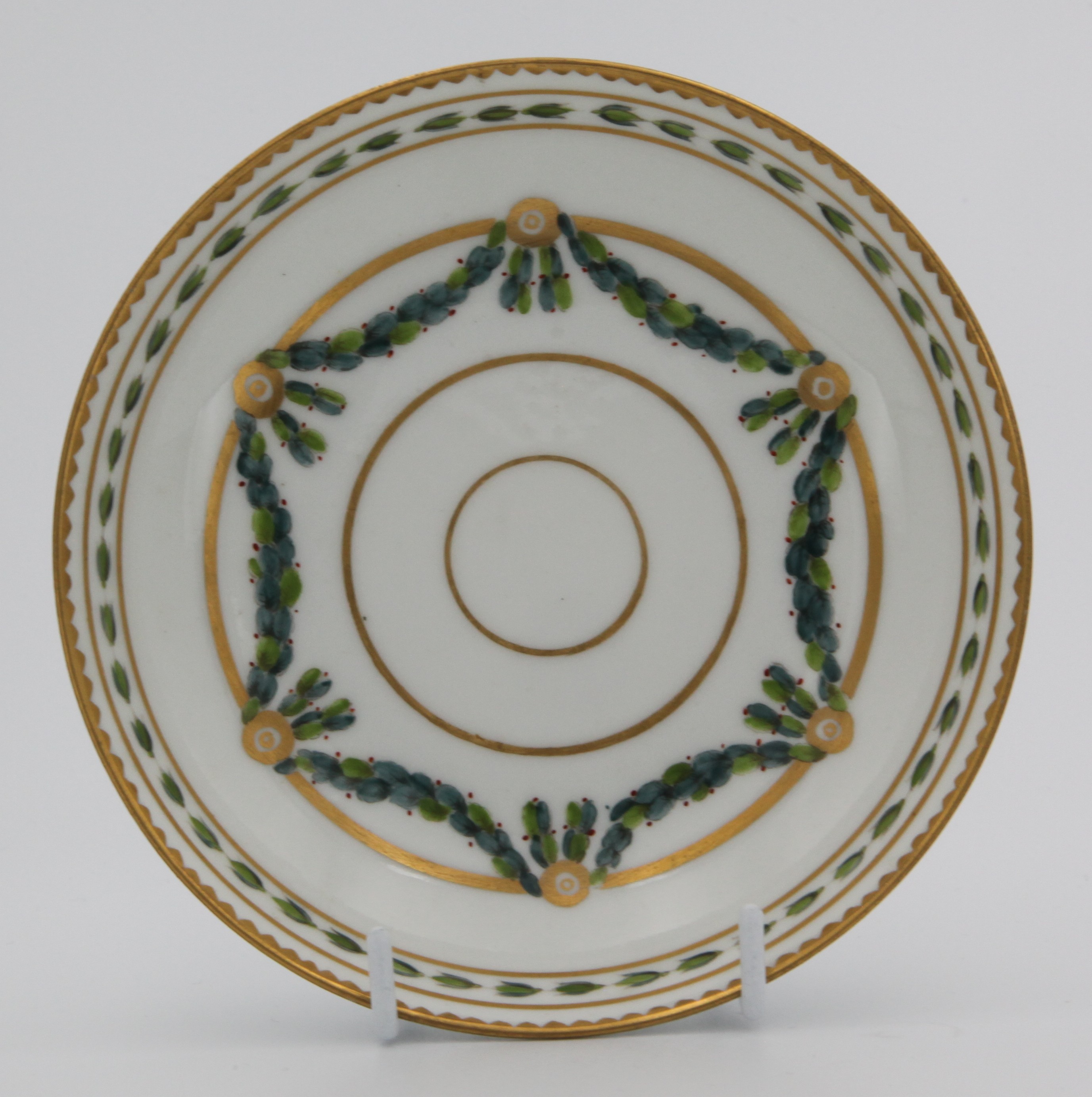 A Bristol coffee saucer, painted in green and iron red with laurel swags, suspended from gilt bands within green leaf scroll bands and gilt dentil rims, circa 1775. Blue X3 mark