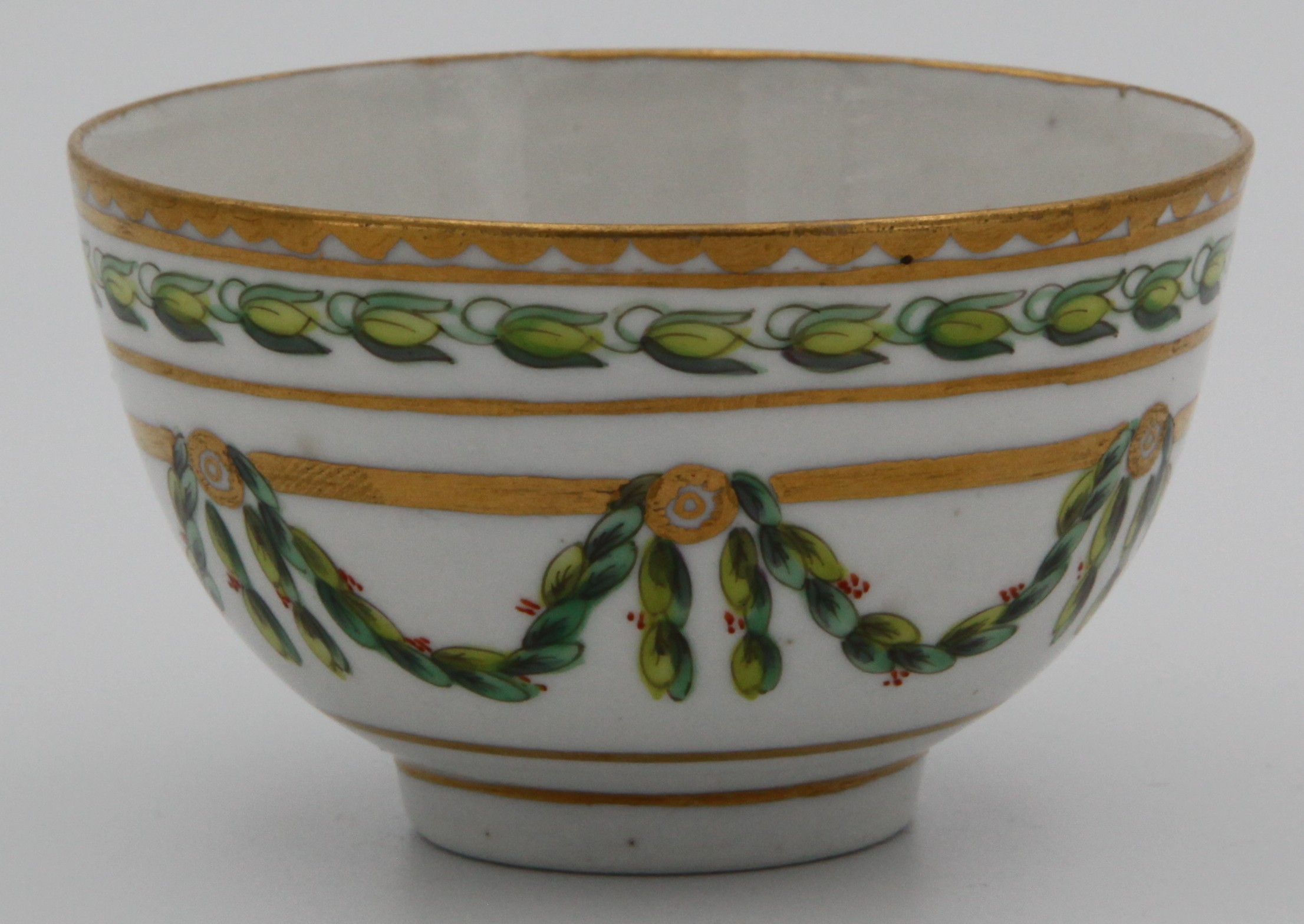A Bristol teabowl, painted in green and iron red with laurel swags, suspended from gilt flower head roundels mounted on a heavy gilt band, within stiff leaf and gilt dentil borders, blue X1. mark