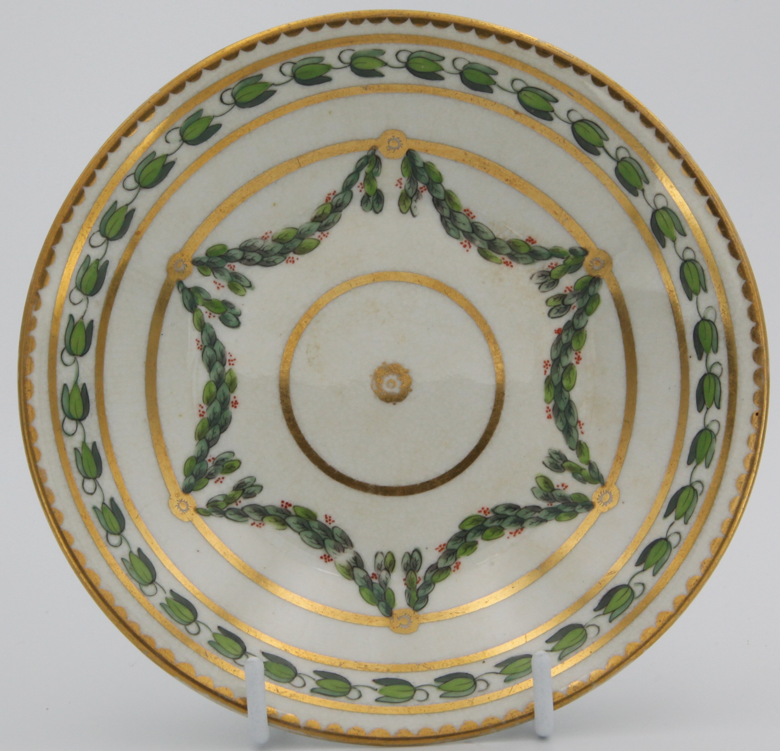 A Bristol-style coffee saucer, painted in green and iron red with laurel swags, suspended from gilt bands within green leaf scroll bands and gilt dentil rims. Mark: red crown symbolsr. Crazing on saucer and marks indicate replacement.