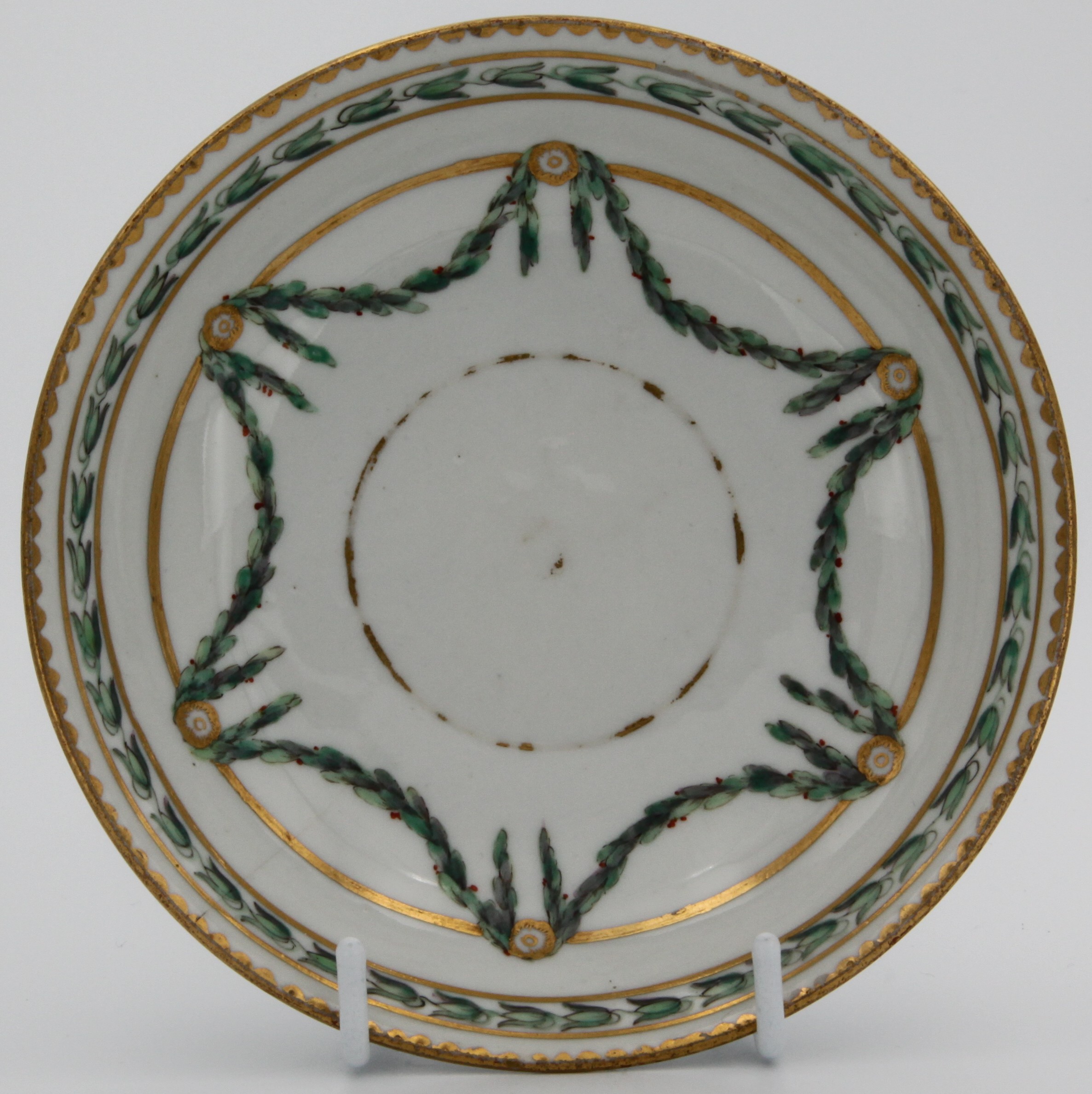 A Bristol coffee saucer, painted in green and iron red with laurel swags, suspended from gilt bands within green leaf scroll bands and gilt dentil rims, circa 1775. Blue X gilt 5. mark