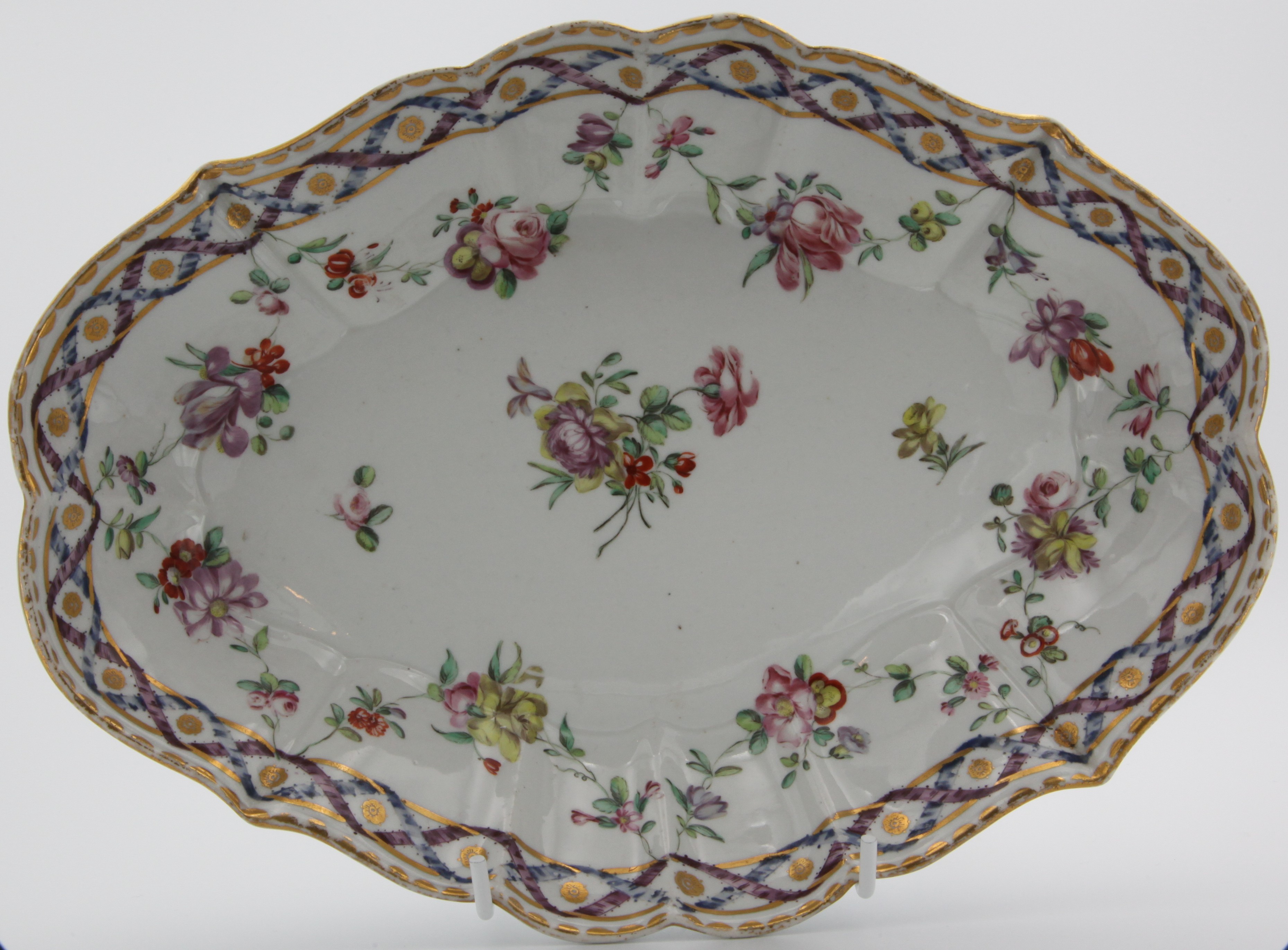 A Bristol fluted lozenge-shaped dish, painted in colourful enamels with a flower spray and scattered flowers, the puce and blue interlocked border with gilt flower head medallions, and suspending swags of flowers and leaves, gilt dentil rim, 10" wide, circa 1775, blue painted X mark. Condition: perfect. See Albert Amor, The Alfred Trapnell Collection, 1912, numbers 170-181, for a part dessert service in this pattern