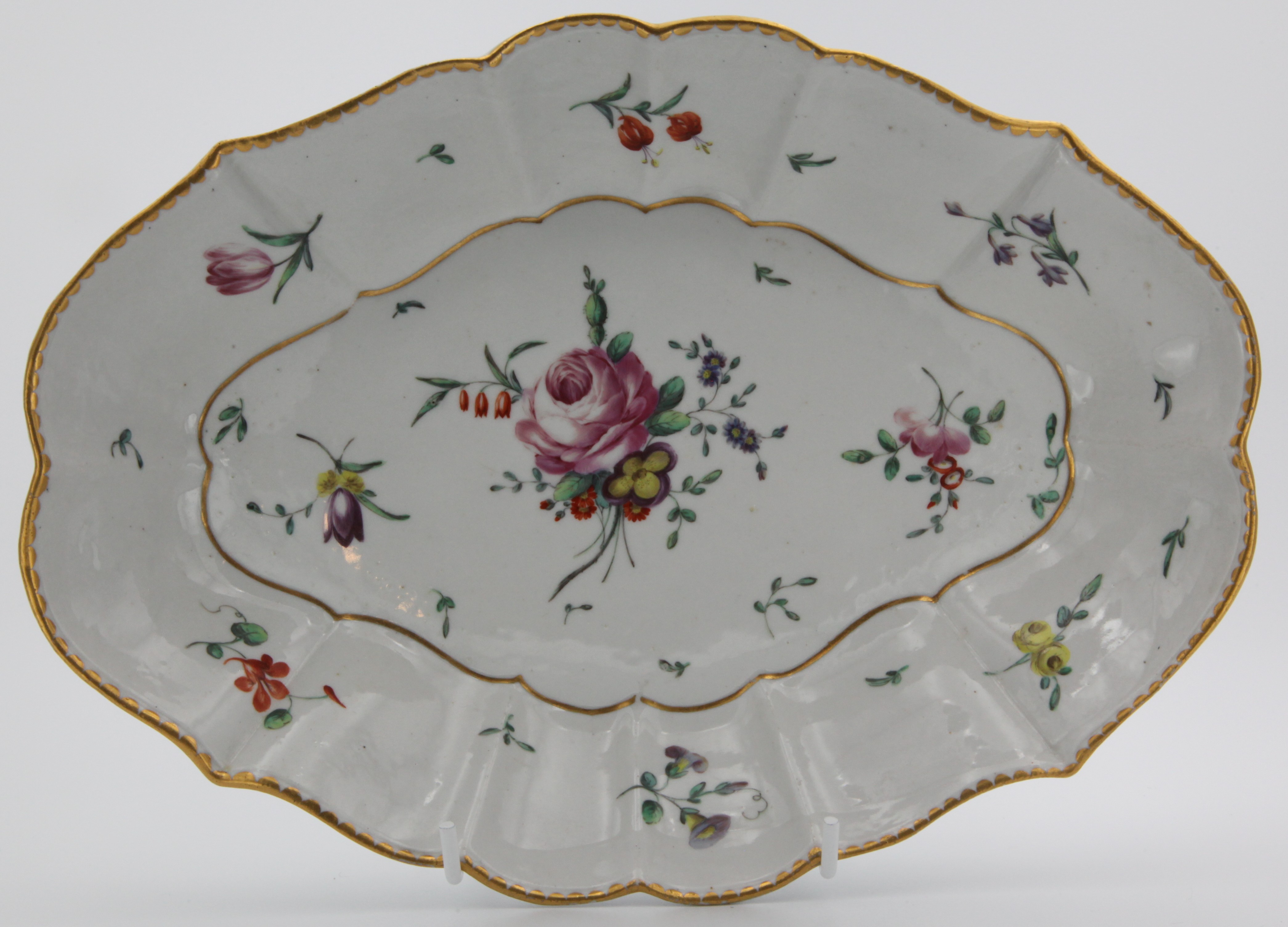 A Bristol fluted lozenge shaped dish, finely painted in coloured enamels with a spray of flowers and leaves, and scattered flowers, within a gilt dentil border, circa 1772-74, blue painted X mark, remnants of rubbed gilt numeral (5?).