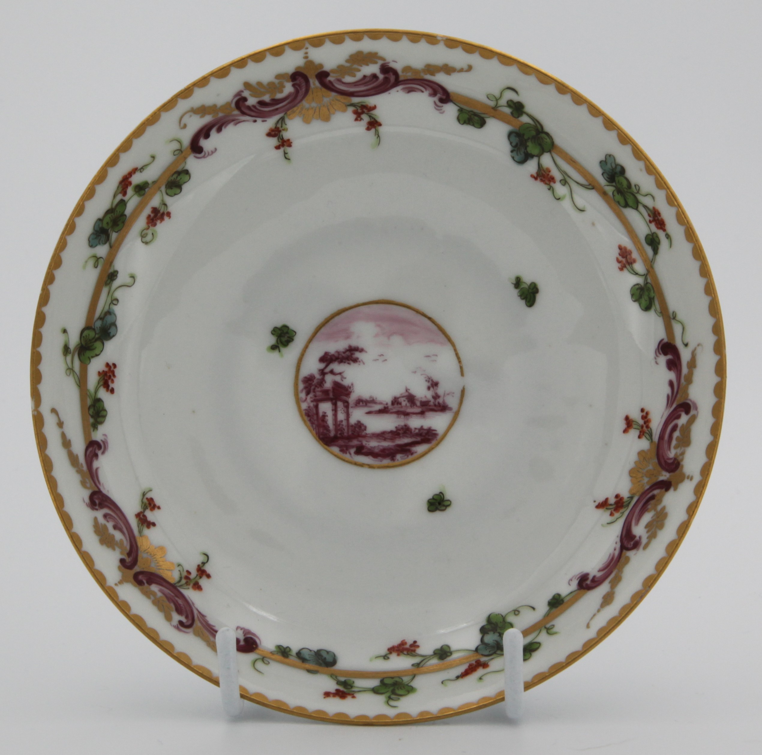 A Bristol tea  saucer, from the Mark Harford Service, each piece painted in purple monochrome with landscape roundels, within a gilt band, the border with iron red flowers and leaves, and puce ‘C’ scrolls, entwined with a gilt band, gilt dentil rims, circa 1772, no marks.
