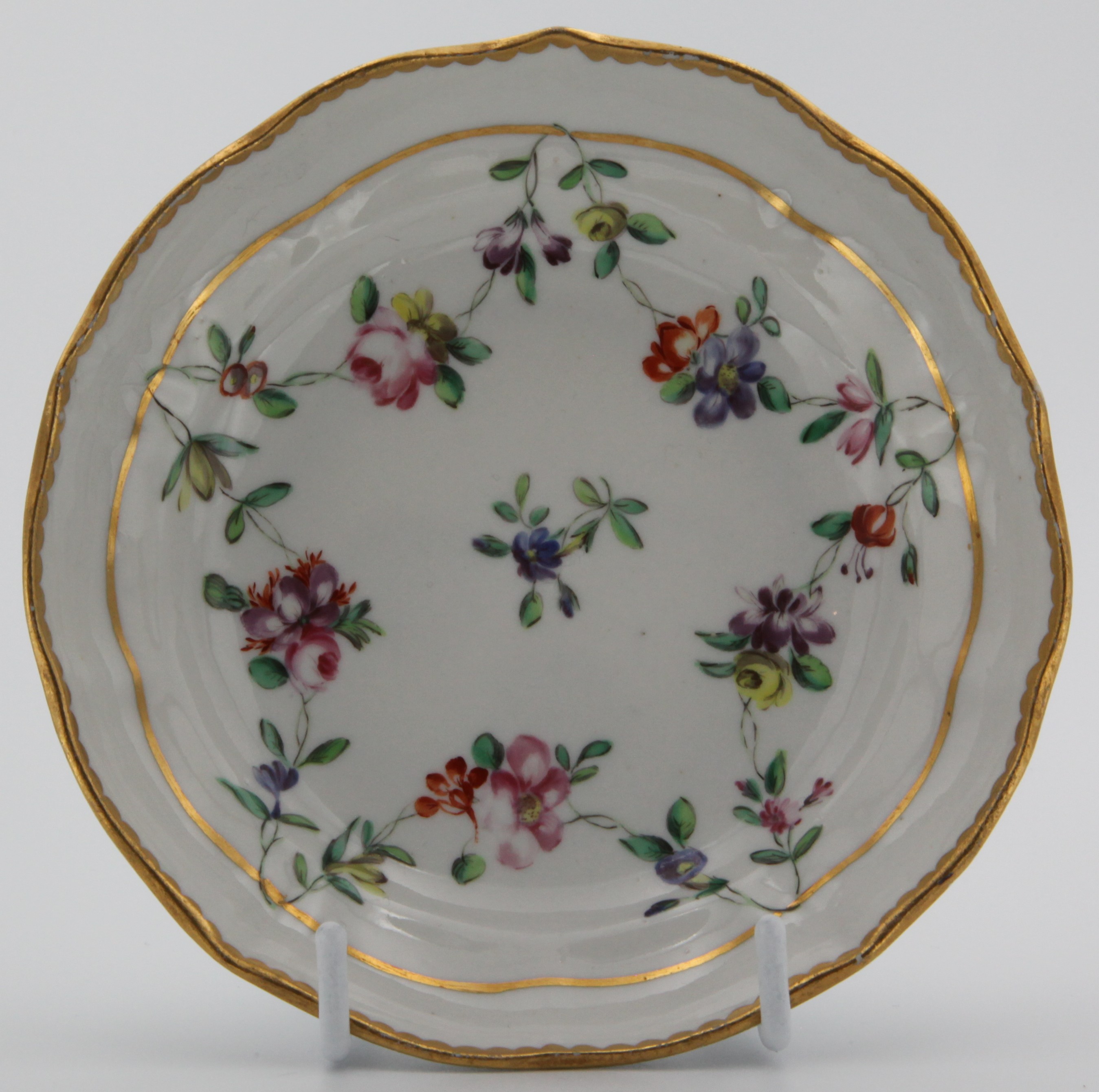 A Bristol ogee shaped coffee saucer, painted in coloured enamels with swags of flowers and leaves, suspended from gilt bands, gilt dentil borders, circa 1775, blue painted X and numeral 5 mark