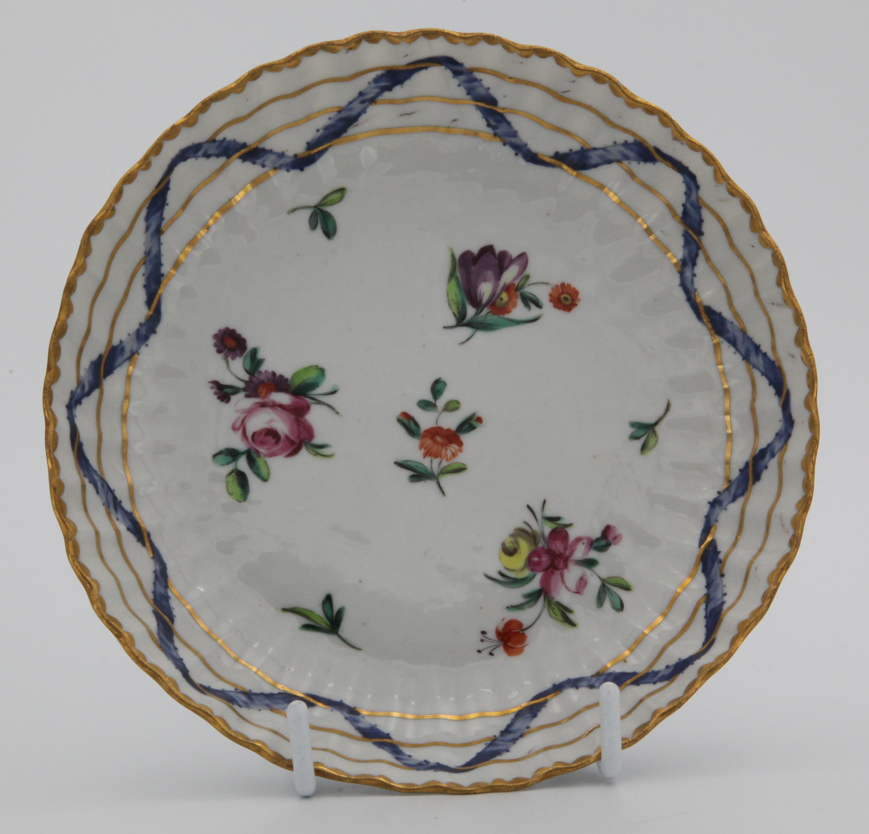 A Bristol ‘Nelson Pattern’ fluted tea saucer, painted in coloured enamels with scattered flowers and leaves, within scrolling blue ribbons, entwined with three gilt bands, gilt dentil rims, circa 1772, blue painted X mark and numeral 5. See Albert Amor, The Trapnell Collection, 1912, numbers 182-189, for pieces from this service, owned by Admiral Lord Nelson, and by descent to Hilaire, Countess Nelson. It was then in the Firbank Collection