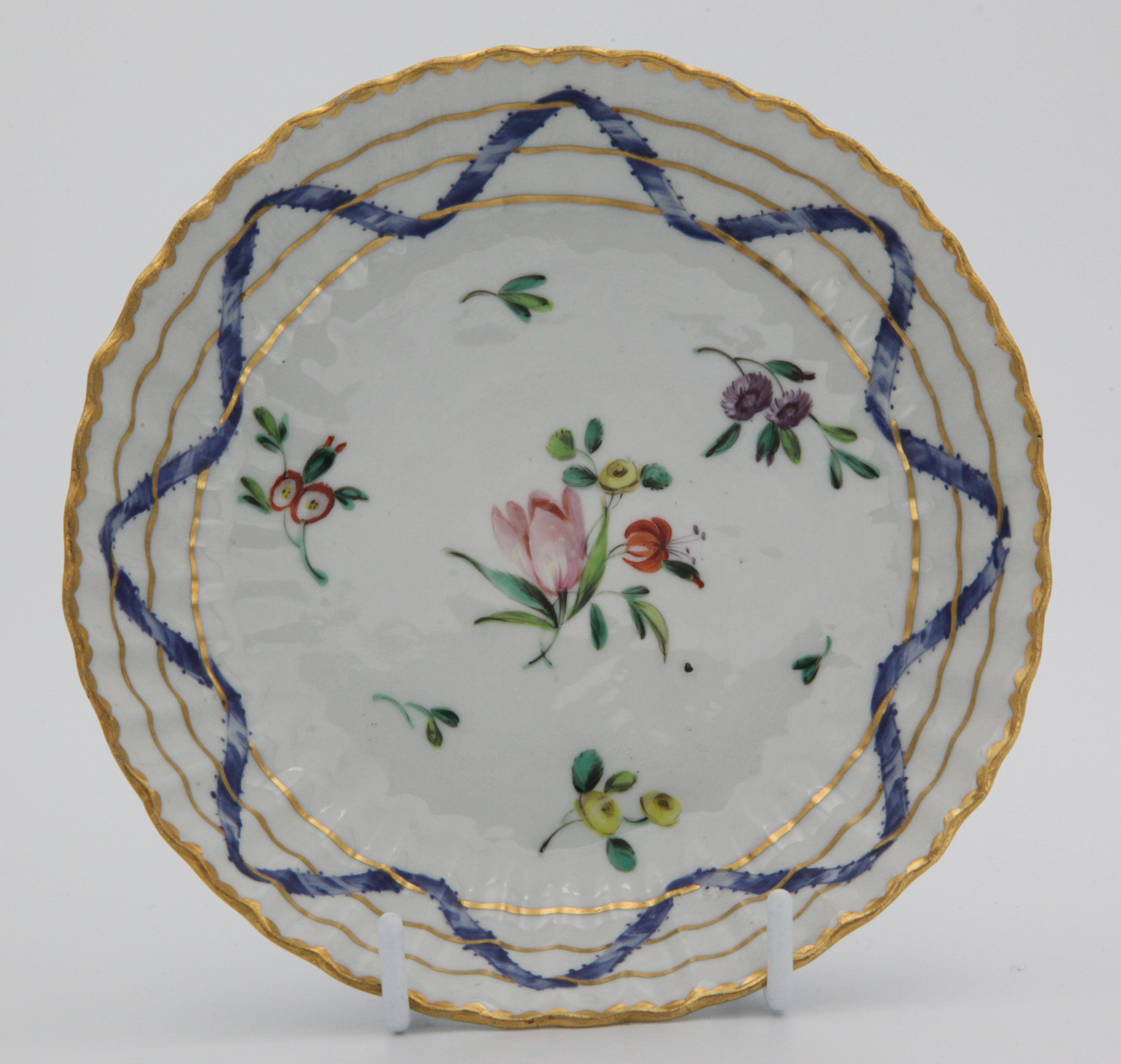 A Bristol ‘Nelson Pattern’ fluted coffee saucer, painted in coloured enamels with scattered flowers and leaves, within scrolling blue ribbons, entwined with three gilt bands, gilt dentil rims, circa 1772, blue painted B mark to both. See Albert Amor, The Trapnell Collection, 1912, numbers 182-189, for pieces from this service,  owned by Admiral Lord Nelson, and by descent to Hilaire, Countess Nelson.