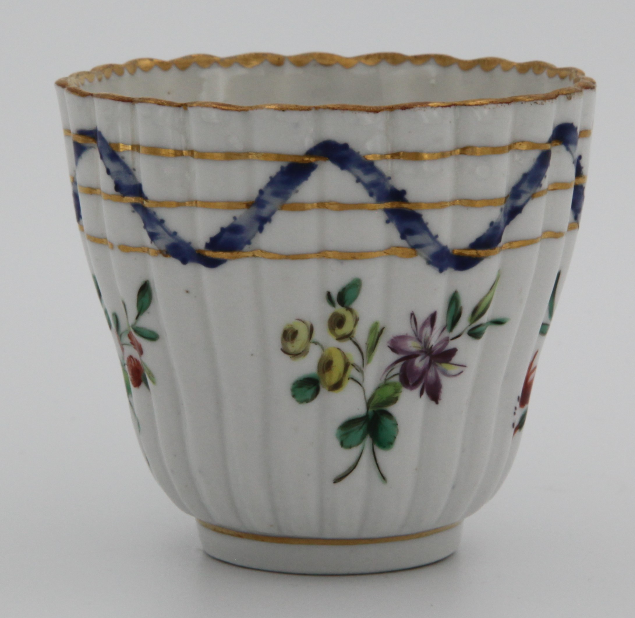 A Bristol ‘Nelson Pattern’ fluted coffee cup with harebell moulded scroll handle, painted in coloured enamels with scattered flowers and leaves, within scrolling blue ribbons, entwined with three gilt bands, gilt dentil rims, circa 1772, blue painted B mark to both. See Albert Amor, The Trapnell Collection, 1912, numbers 182-189, for pieces from this service,  owned by Admiral Lord Nelson, and by descent to Hilaire, Countess Nelson.