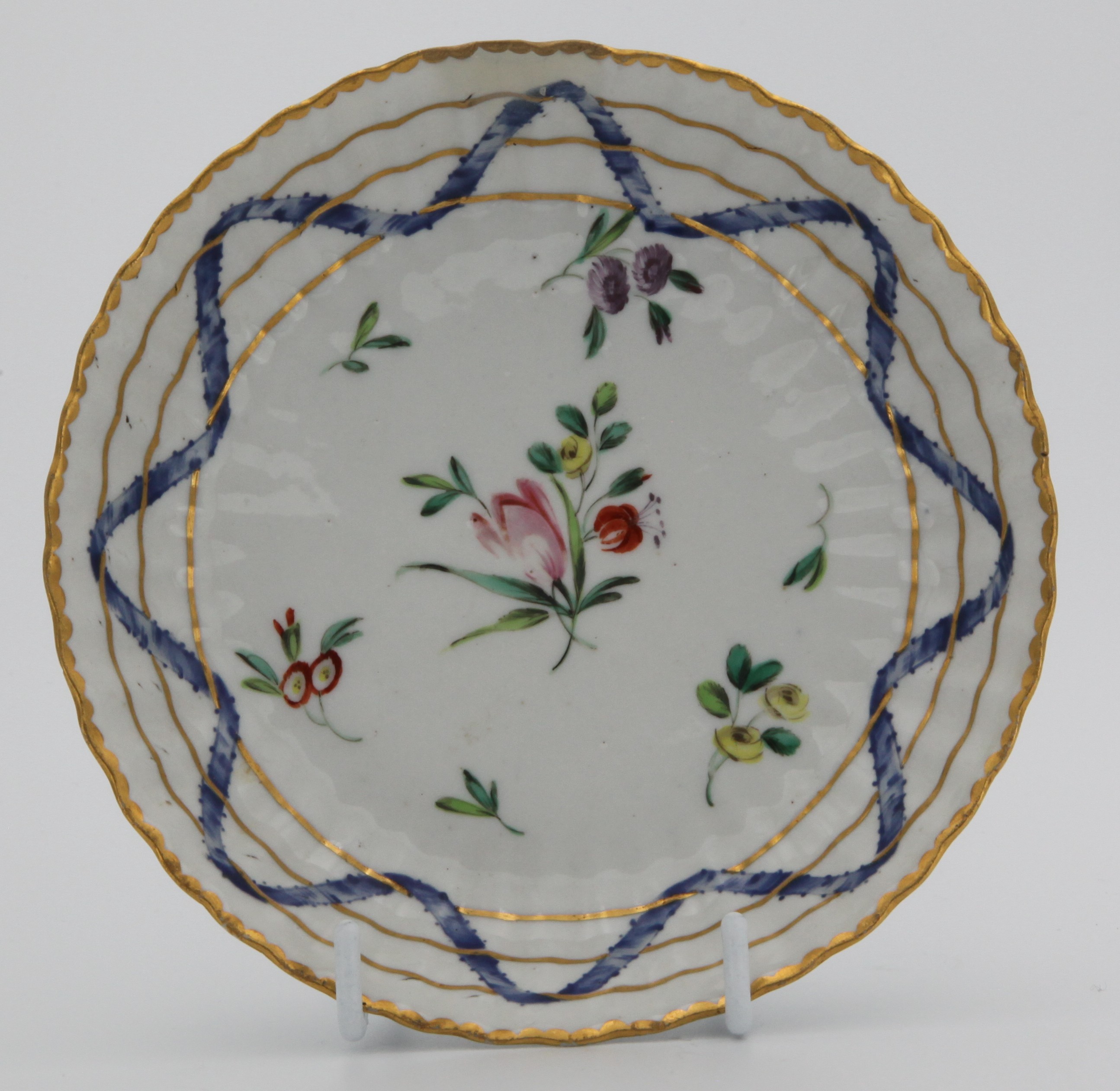 A Bristol ‘Nelson Pattern’ fluted tea saucer, the cup with harebell moulded scroll handle, painted in coloured enamels with scattered flowers and leaves, within scrolling blue ribbons, entwined with three gilt bands, gilt dentil rims, circa 1772, blue painted B mark and numeral 5. See Albert Amor, The Trapnell Collection, 1912, numbers 182-189, for pieces from this service, owned by Admiral Lord Nelson, and by descent to Hilaire, Countess Nelson.