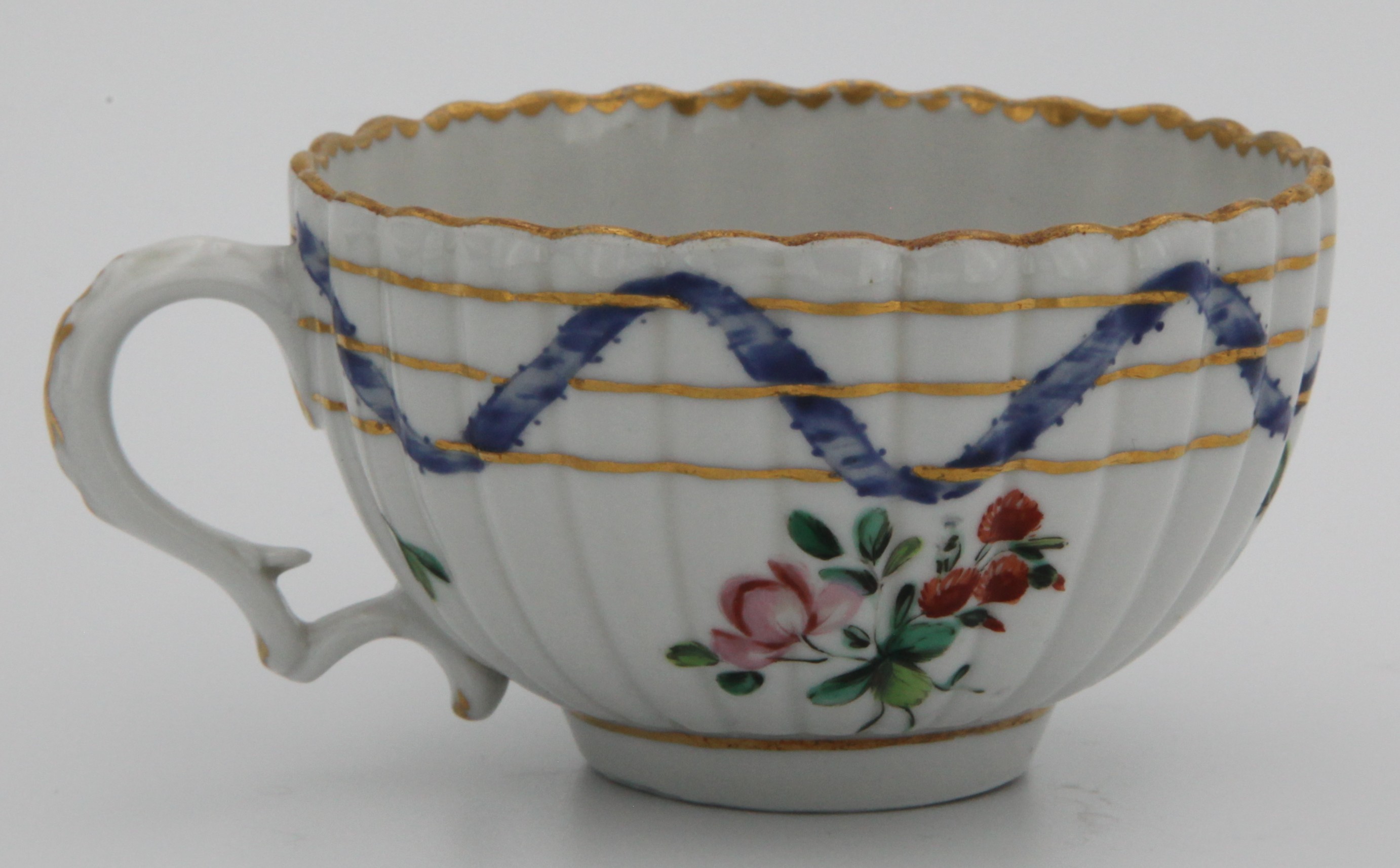A Bristol ‘Nelson Pattern’ fluted teacup with harebell moulded scroll handle, painted in coloured enamels with scattered flowers and leaves, within scrolling blue ribbons, entwined with three gilt bands, gilt dentil rims, circa 1772, blue painted B mark and numeral 5. See Albert Amor, The Trapnell Collection, 1912, numbers 182-189, for pieces from this service, owned by Admiral Lord Nelson, and by descent to Hilaire, Countess Nelson.