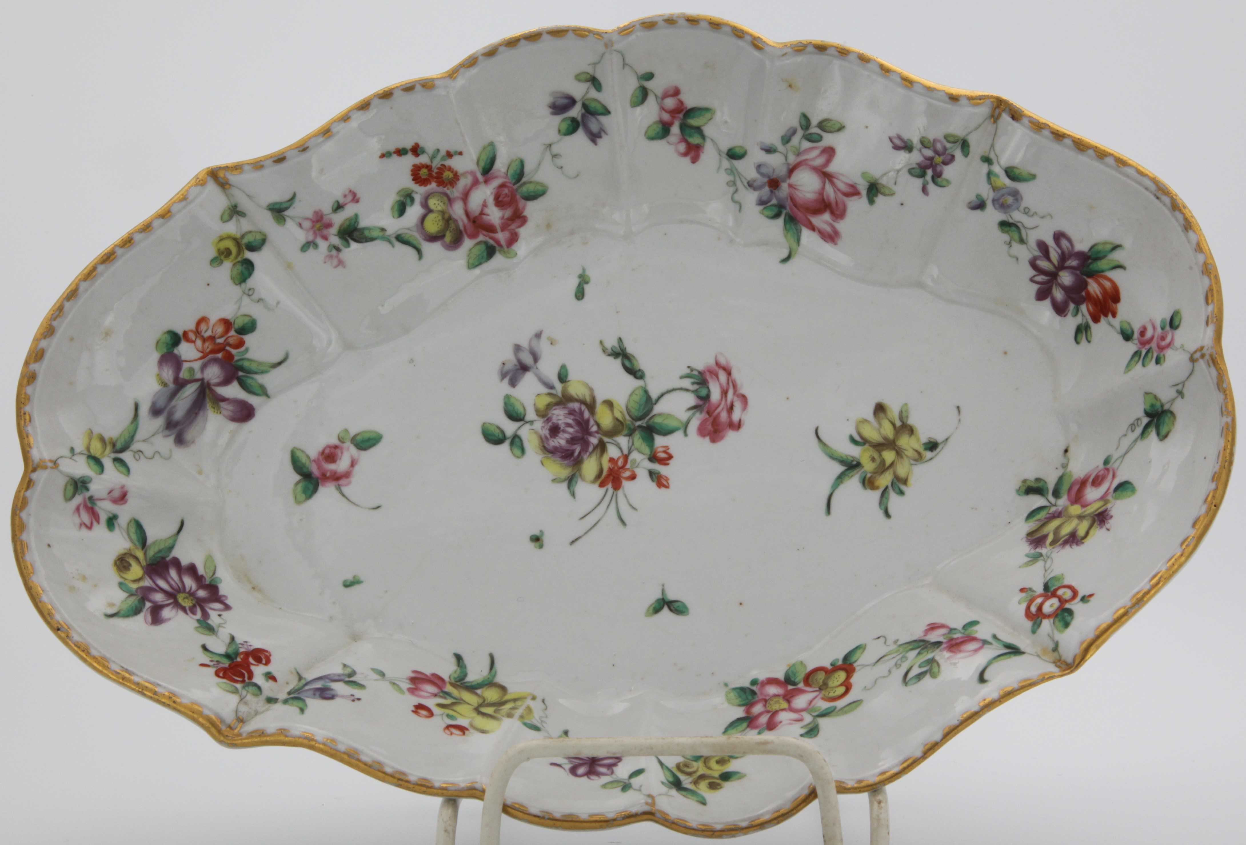 A fluted lozenge-shaped Bristol dish painted with colourful garlands of flowers and leaves suspended from gilt loops on gilt dentil rim and with scattered flowers, 25 1/2cm, blue X mark