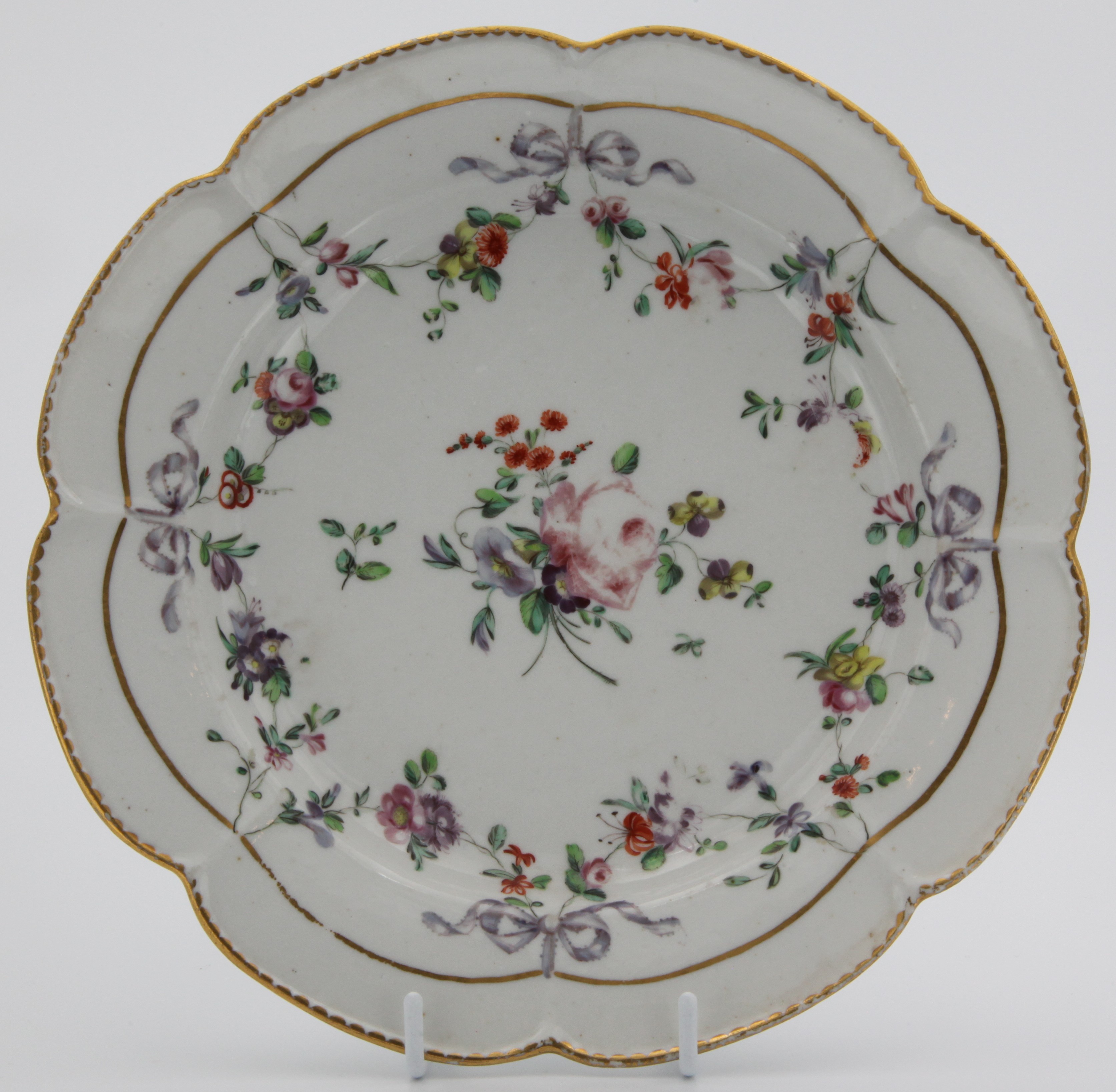 A lobed Bristol plate, painted in coloured enamels with garlands of flowers and leaves, suspended from purple bows and hanging from a gilt line, and with central flower spray, all surrounded by gilt dentil rim. Blue Xb mark