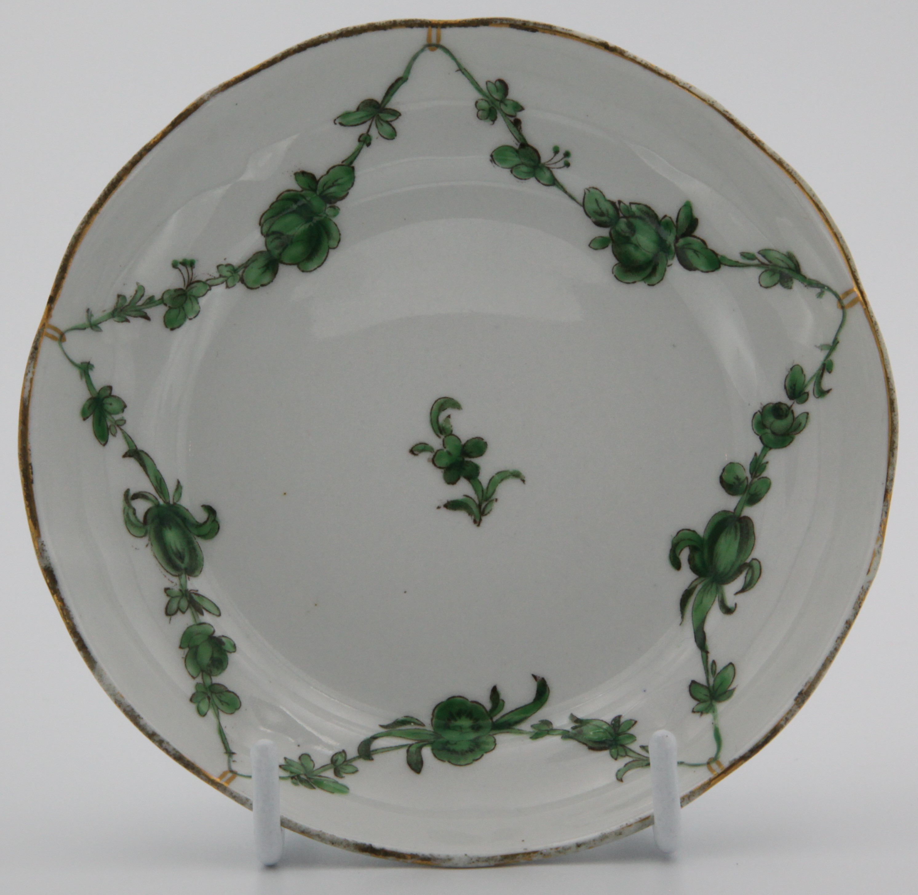 A Bristol lobed, ogee-shaped tea saucer painted with green monochrome garlands of flowers and leaves suspended from gilt loops on gilt rim.  No mark