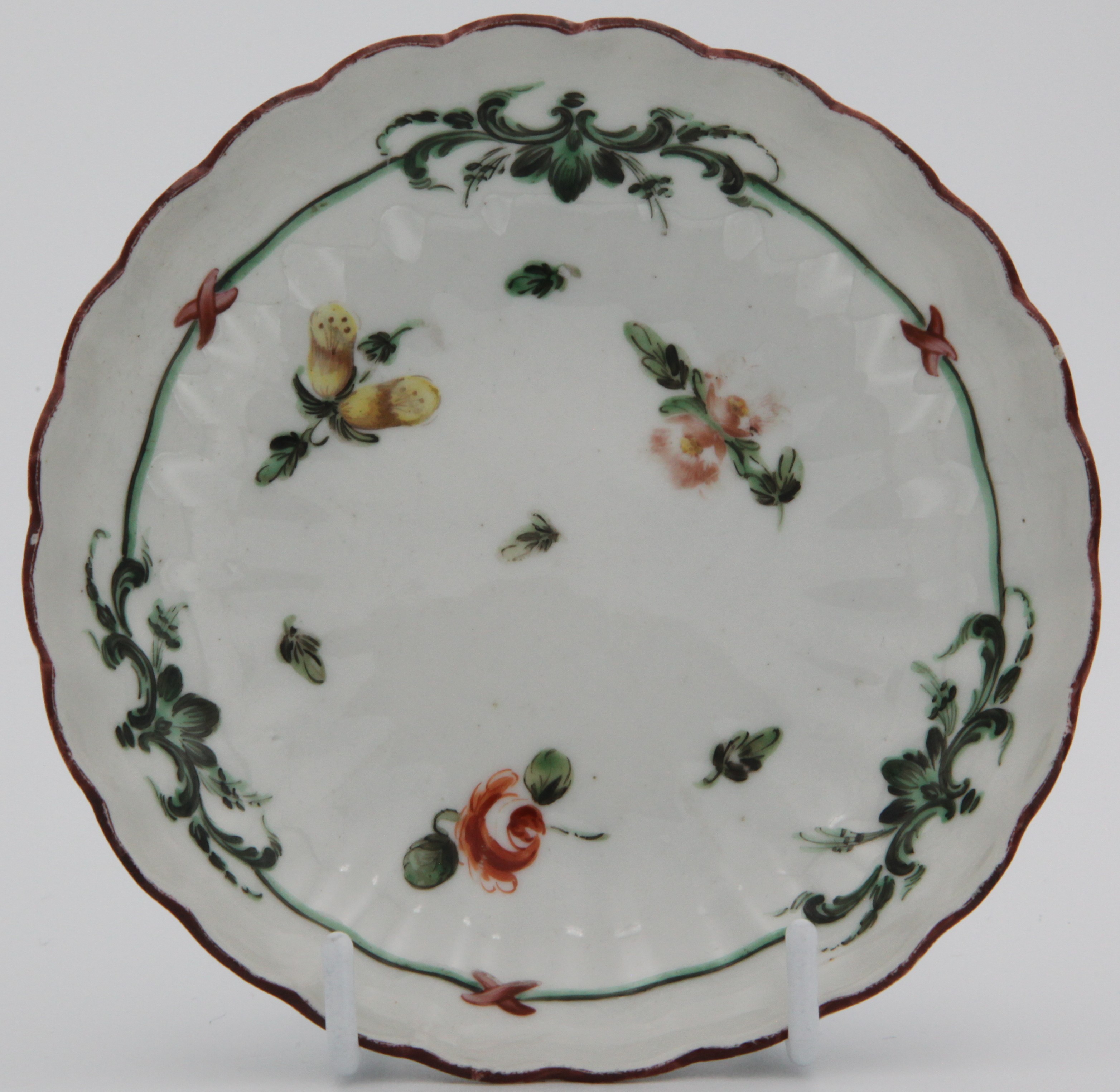 A Bristol fluted saucer painted with green and black band with red ribbons surrounding scattered flowers and leaves, under an iron red rim line. blue X6. mark