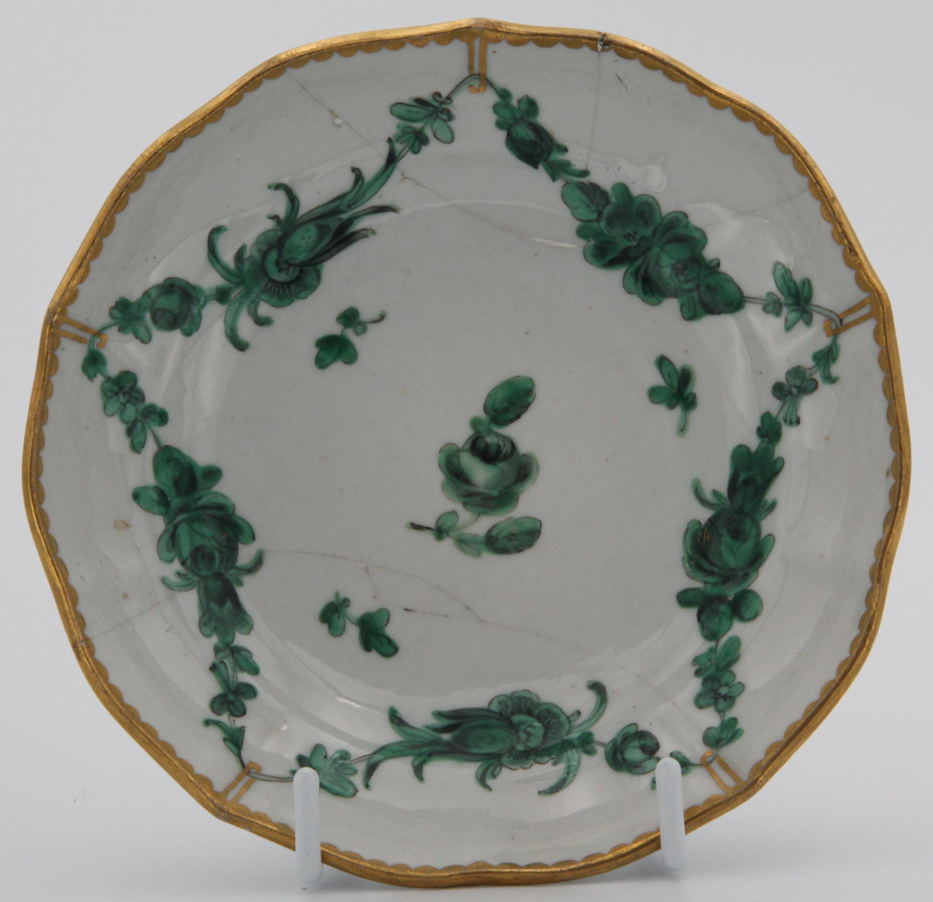 A Bristol slightly lobed ogee-shaped tea saucer, painted with green monochrome garlands suspended from gilt loops hanging from a heavy gilt dentil rim.