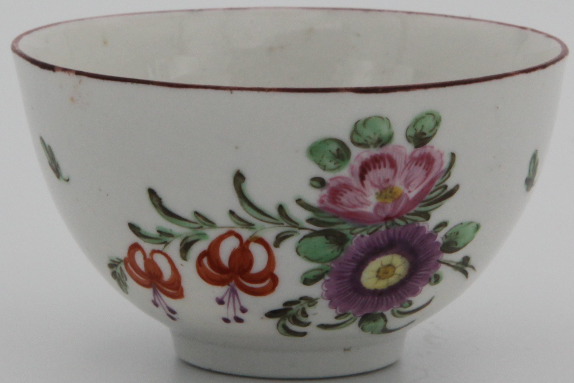 A Bristol tea bowl, painted with a spray of purple, pink and two orange flowers, the reverse with scattered flowers and leaves, a small flower to the base of the bowl, under an iron red line rim. Circa 1775. Mark: painted blue X8.