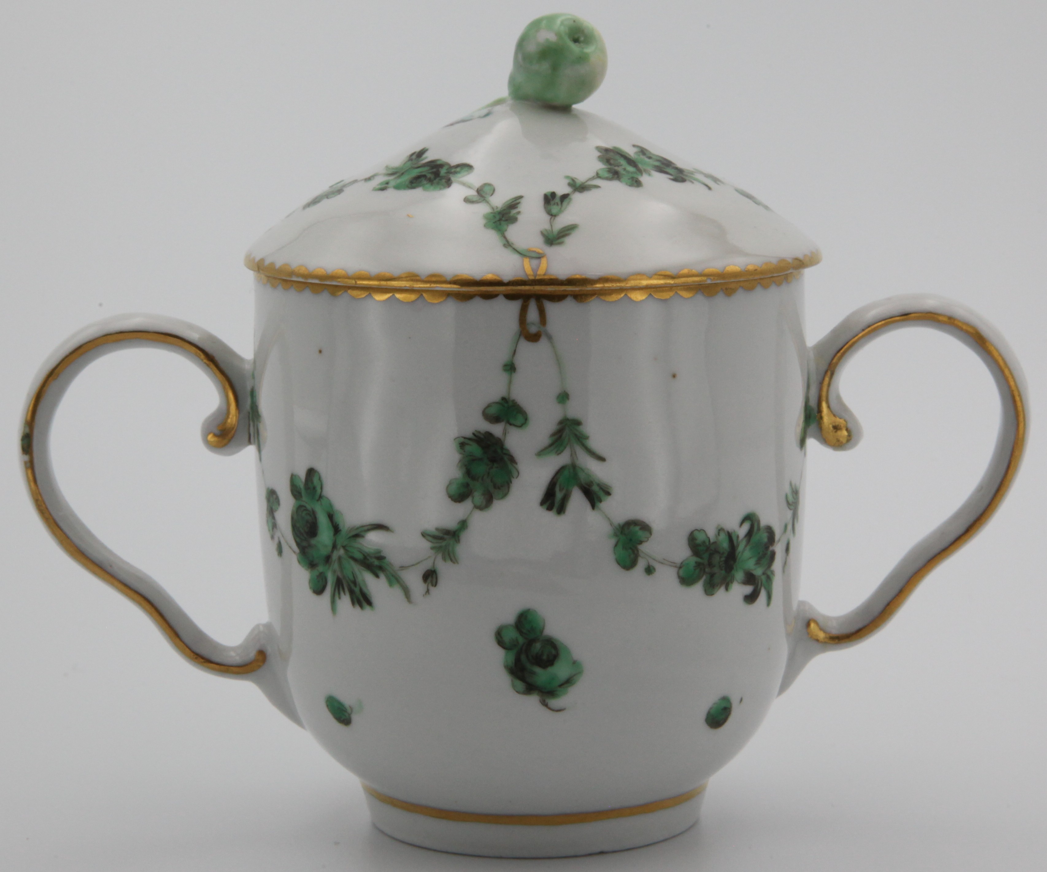 A Bristol chocolate cup and cover, with unusual fruit knop, the cup with two ear shaped handles, painted in green monochrome with swags of flowers and leaves, suspended from gilt loops, gilt dentil and line border, circa 1775, blue painted X and gilt 7. See Frank Hurlbutt, Bristol Porcelain, plate 46, for another example of this form.