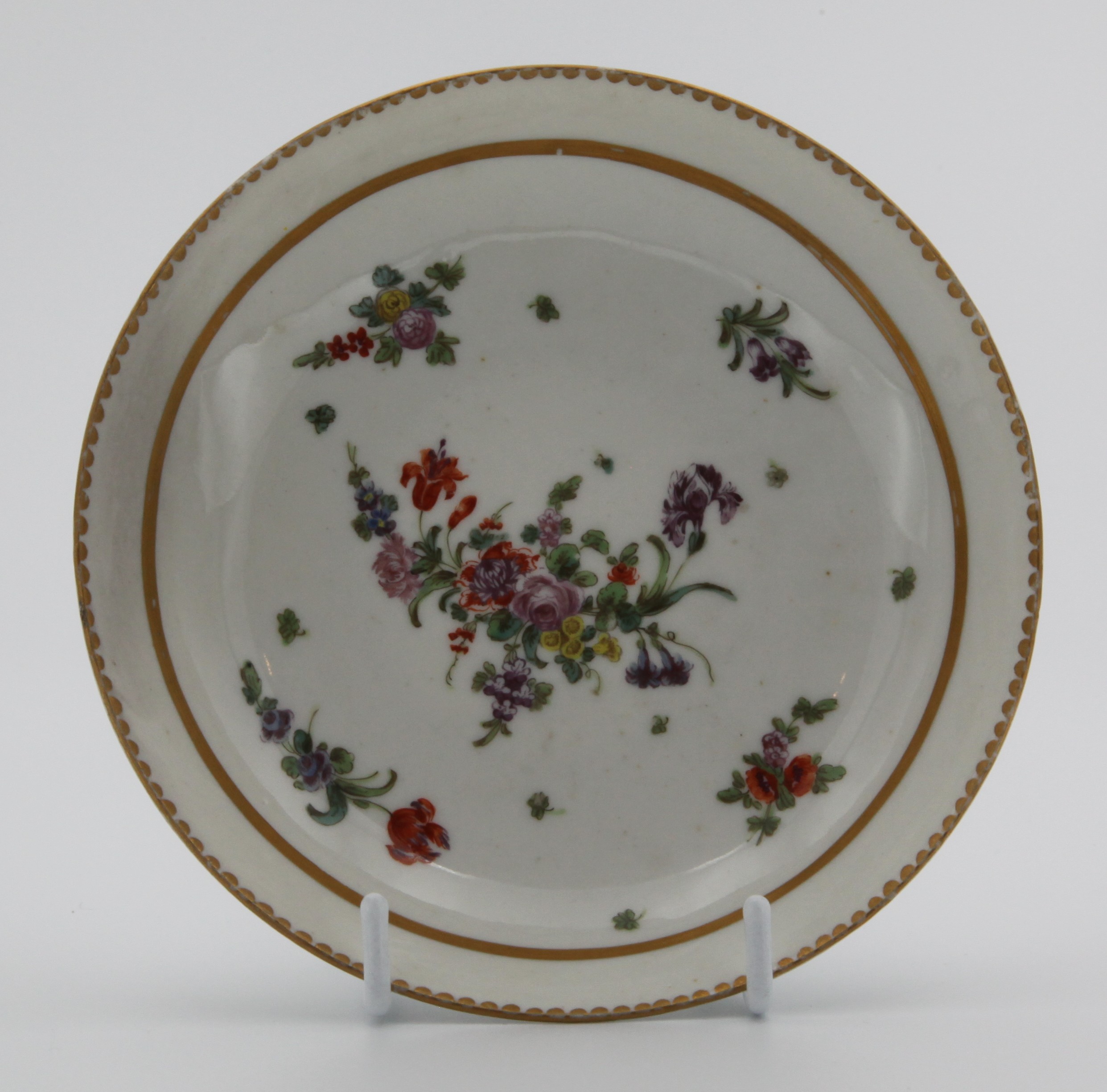 A Bristol saucer painted with a central spray and scattered sprigs under a solid gold border  and a dentil border c. 1773-76.  Mark: none. Has been matched with 1357
