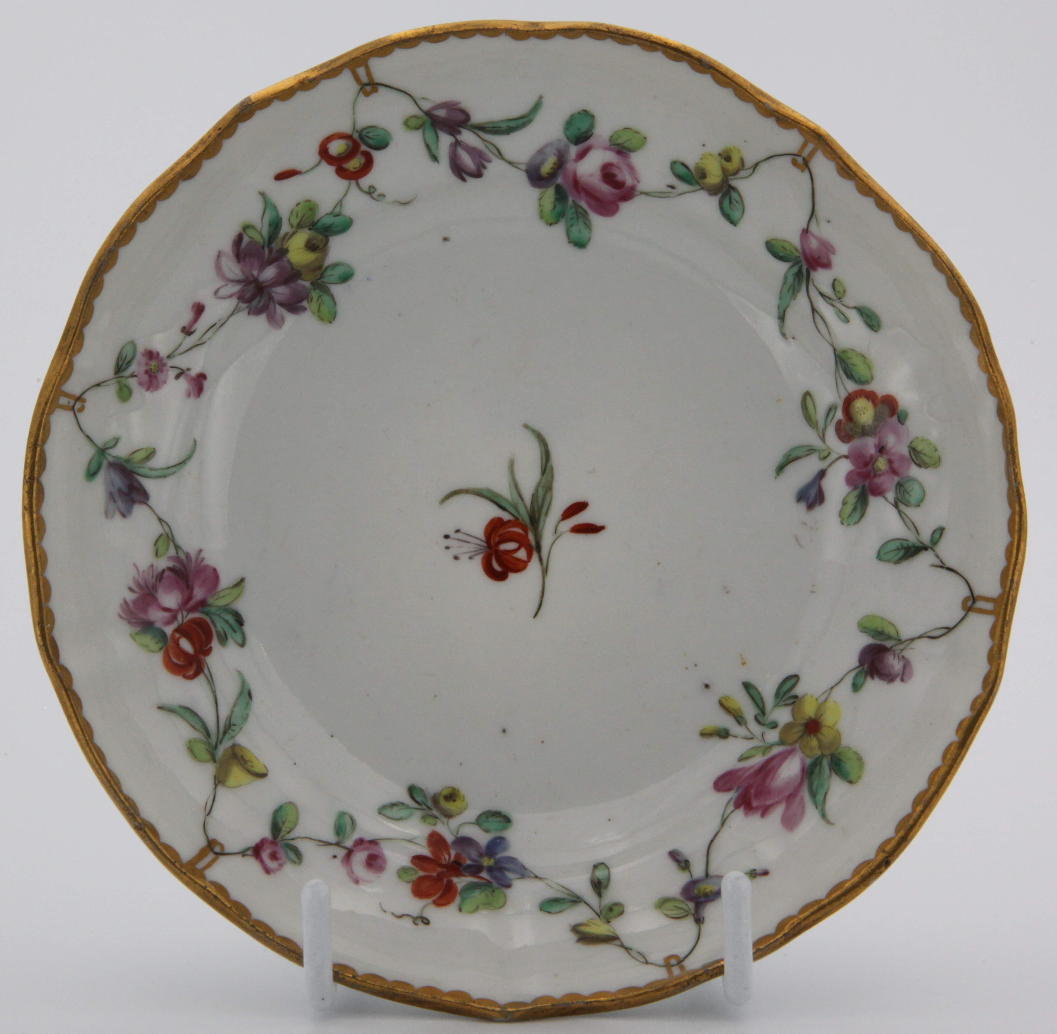 A Bristol saucer painted with chains of flowers hanging from gold loops around a central red flower spray and the loops suspended from a heavy gilt dentil rim. c. 1775-80 Mark: blue Xb.