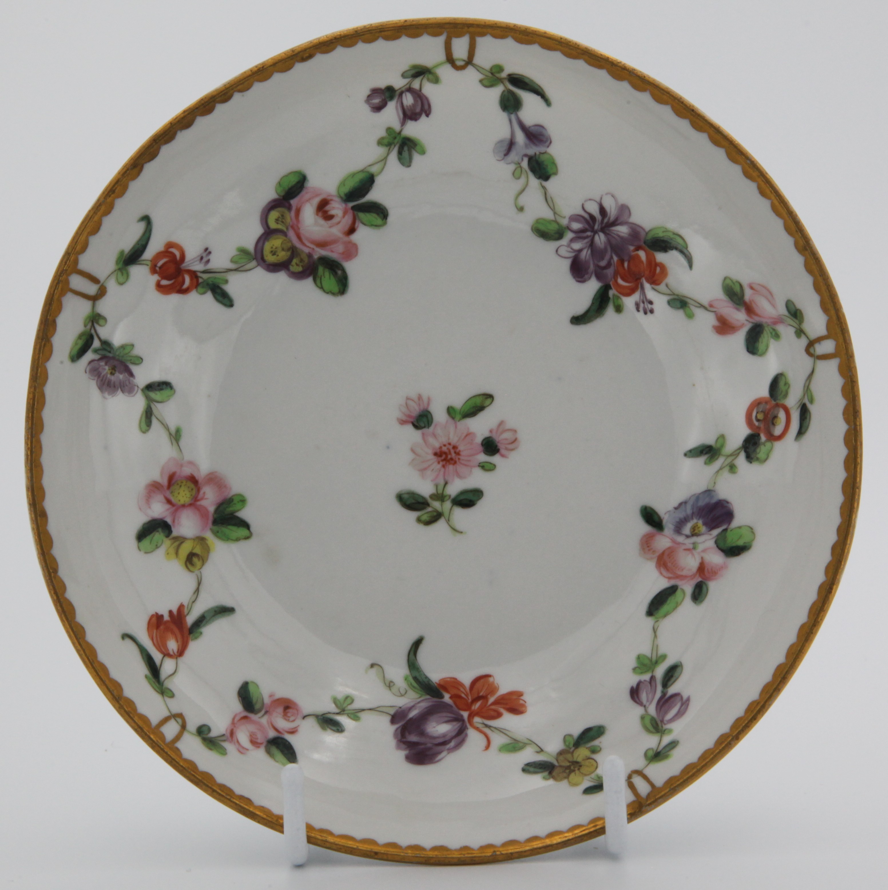 A Bristol saucer painted with chains of flowers hanging from gold loops around a central red flower spray and the loops suspended from a heavy gilt dentil rim. c. 1775-80. Mark: X1.