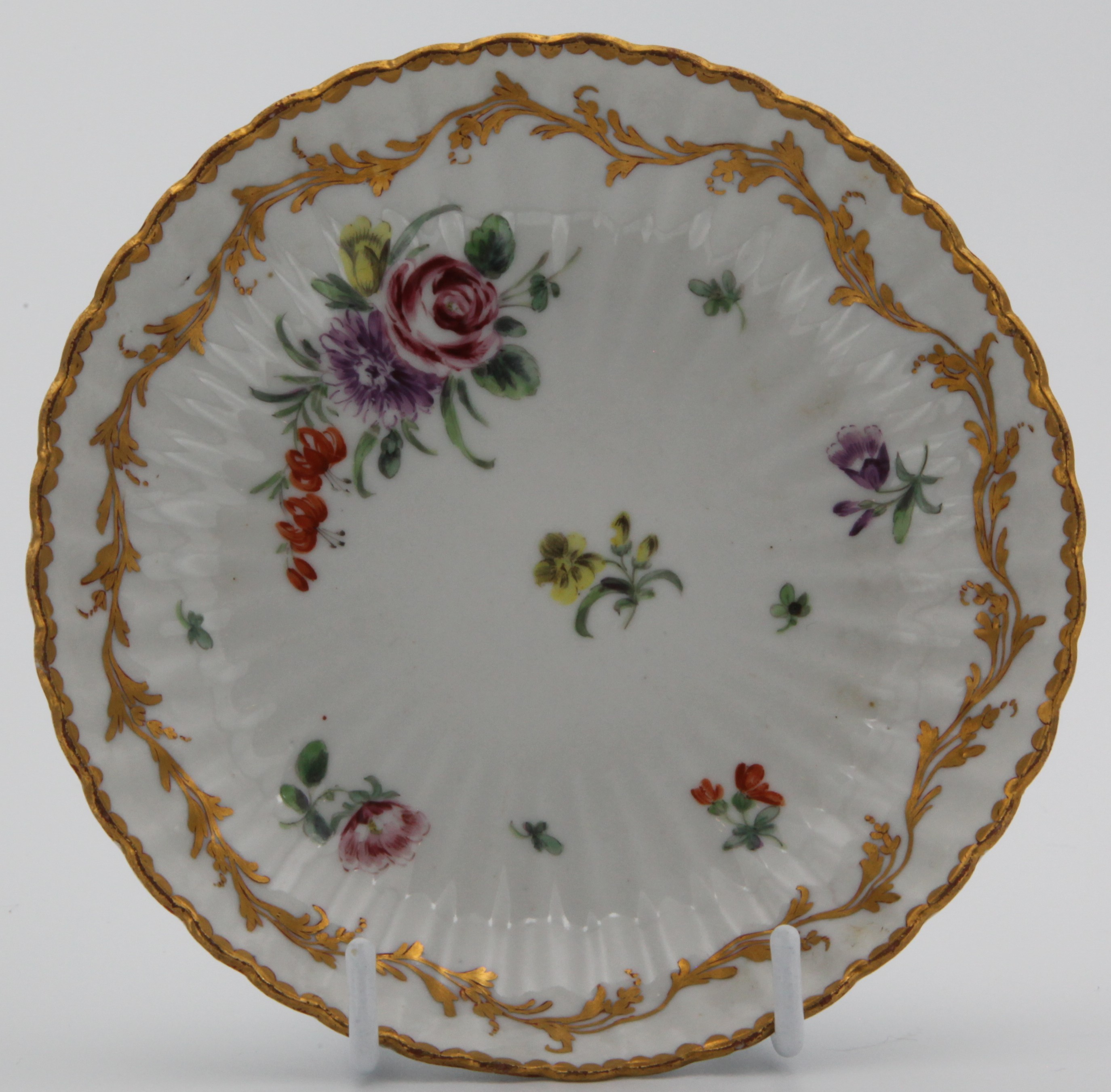 A Bristol fluted saucer painted in colourful enamels with floral sprays and scattered sprigs, an elaborate leafy gilt scrolled border below the gilt dentil rim, blue X gilt 1 mark, c.1775-78. Has been paired with 1356