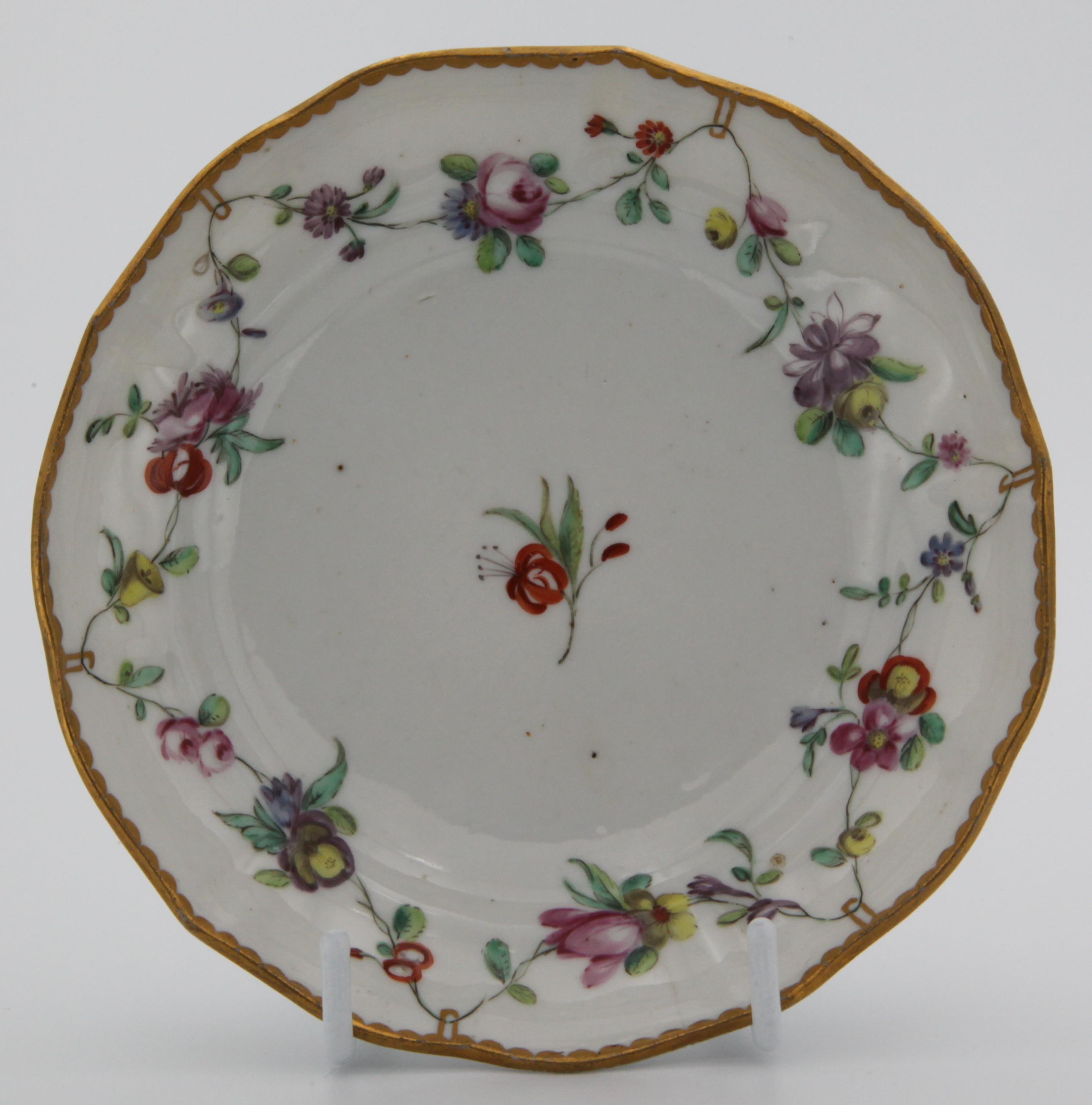 A Bristol saucer painted with chains of flowers hanging from gold loops around a central red flower spray and the loops suspended from a heavy gilt dentil rim. c. 1775-80 Mark: blue Xb.